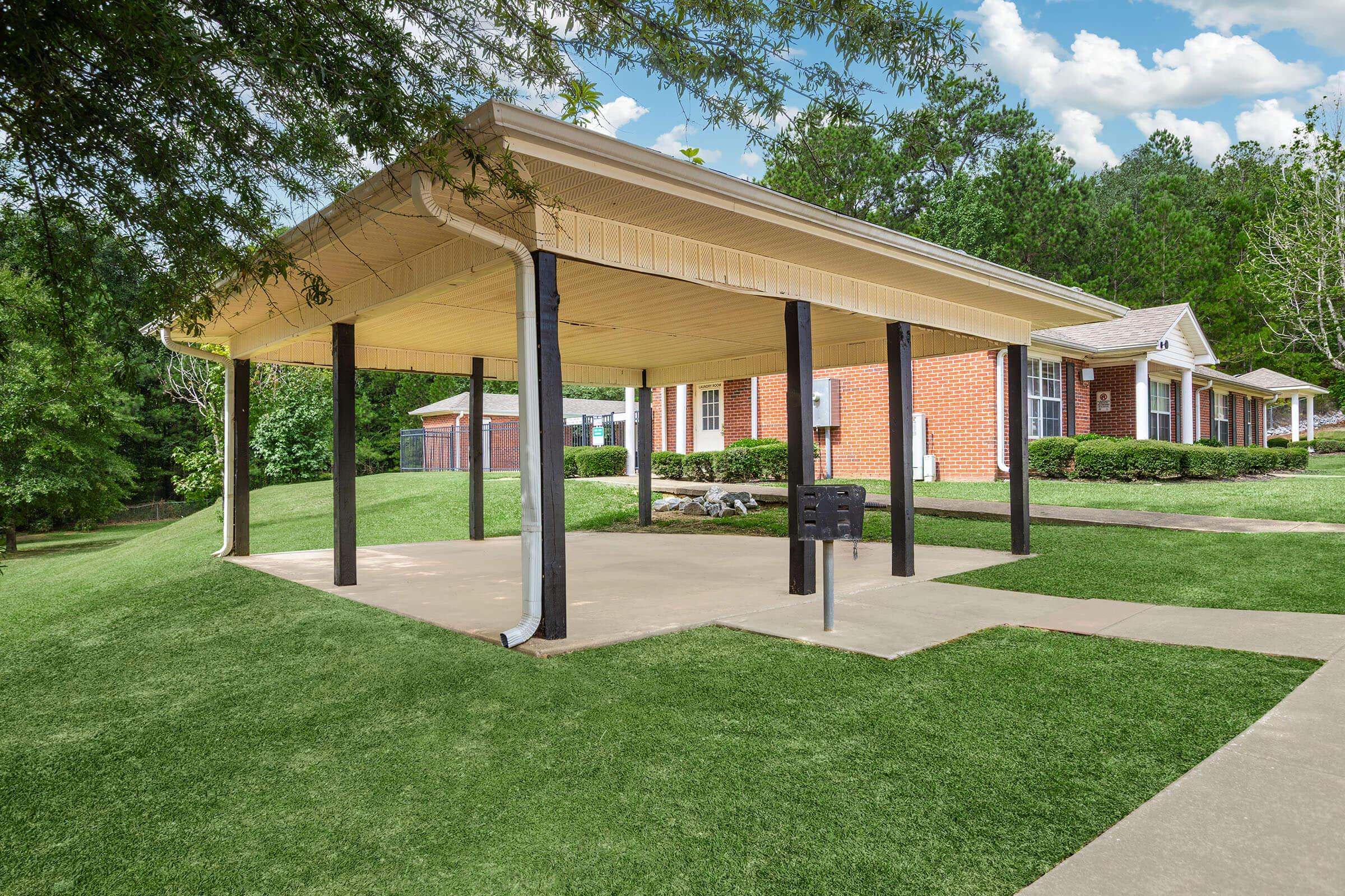 A spacious covered outdoor pavilion with a wooden ceiling and black support beams, situated on a neatly landscaped lawn. In the background, red brick buildings can be seen, surrounded by lush green trees and grass, under a partly cloudy sky.