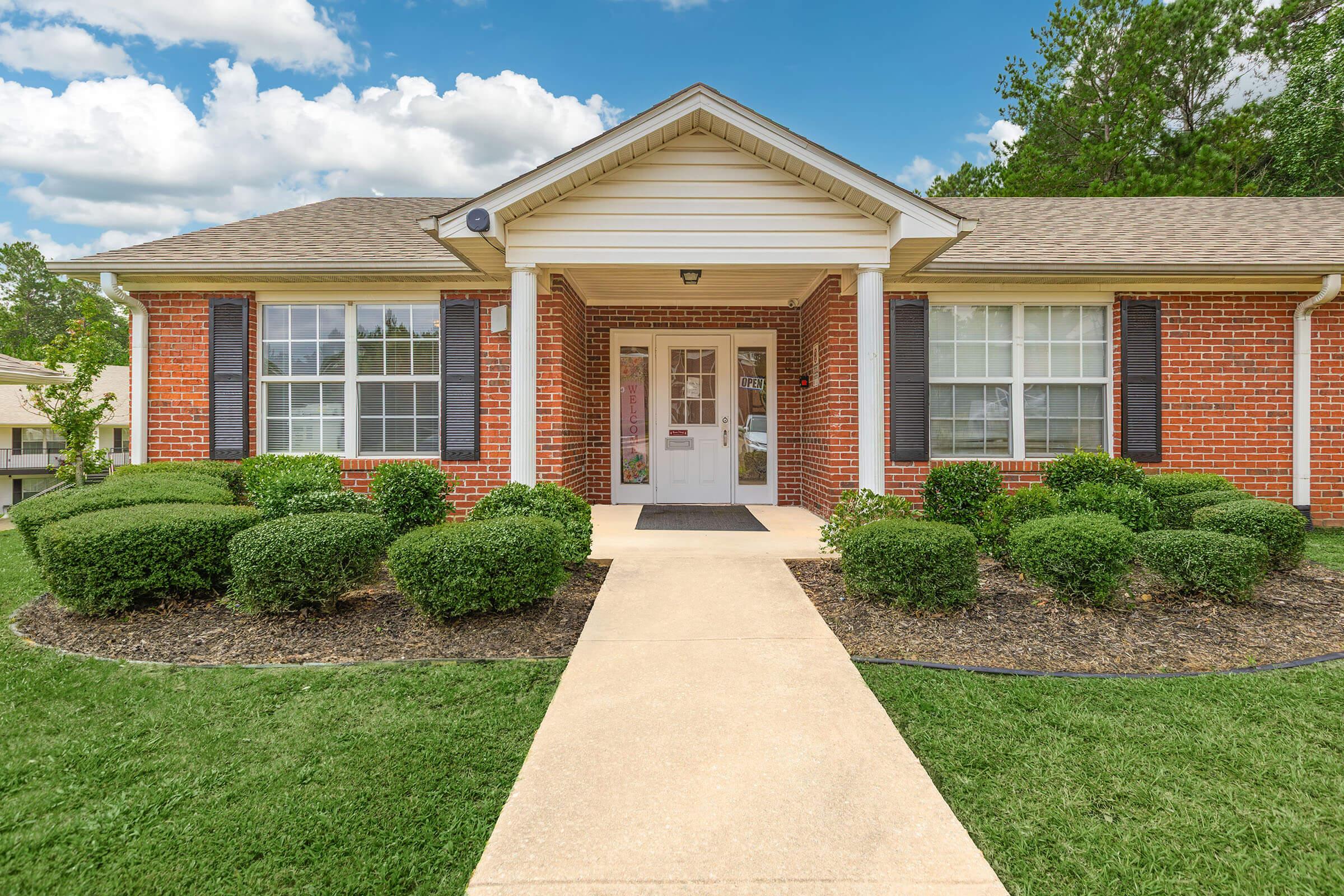 A well-maintained brick building with a welcoming entrance, featuring a concrete walkway leading to the front door. The landscaping includes neatly trimmed bushes and a green lawn under a partly cloudy sky.