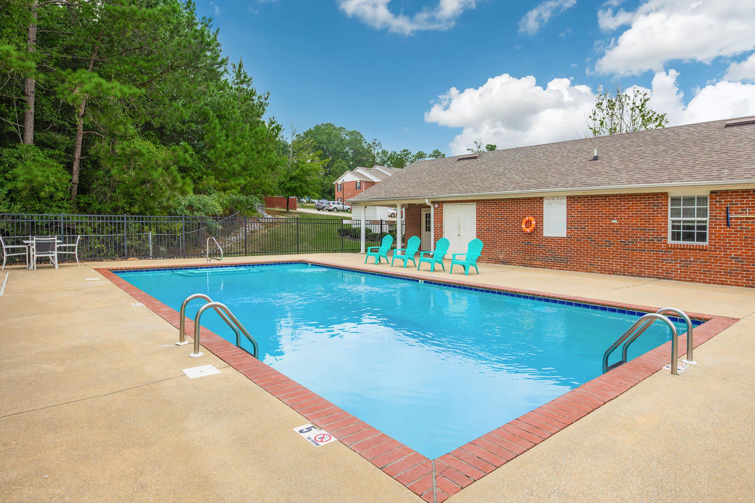 A bright, inviting swimming pool surrounded by a concrete patio. There are several turquoise chairs arranged near the pool. Lush green trees provide some shade in the background, and a brick building with a roof is visible nearby. The sky is partly cloudy, adding to the serene atmosphere.