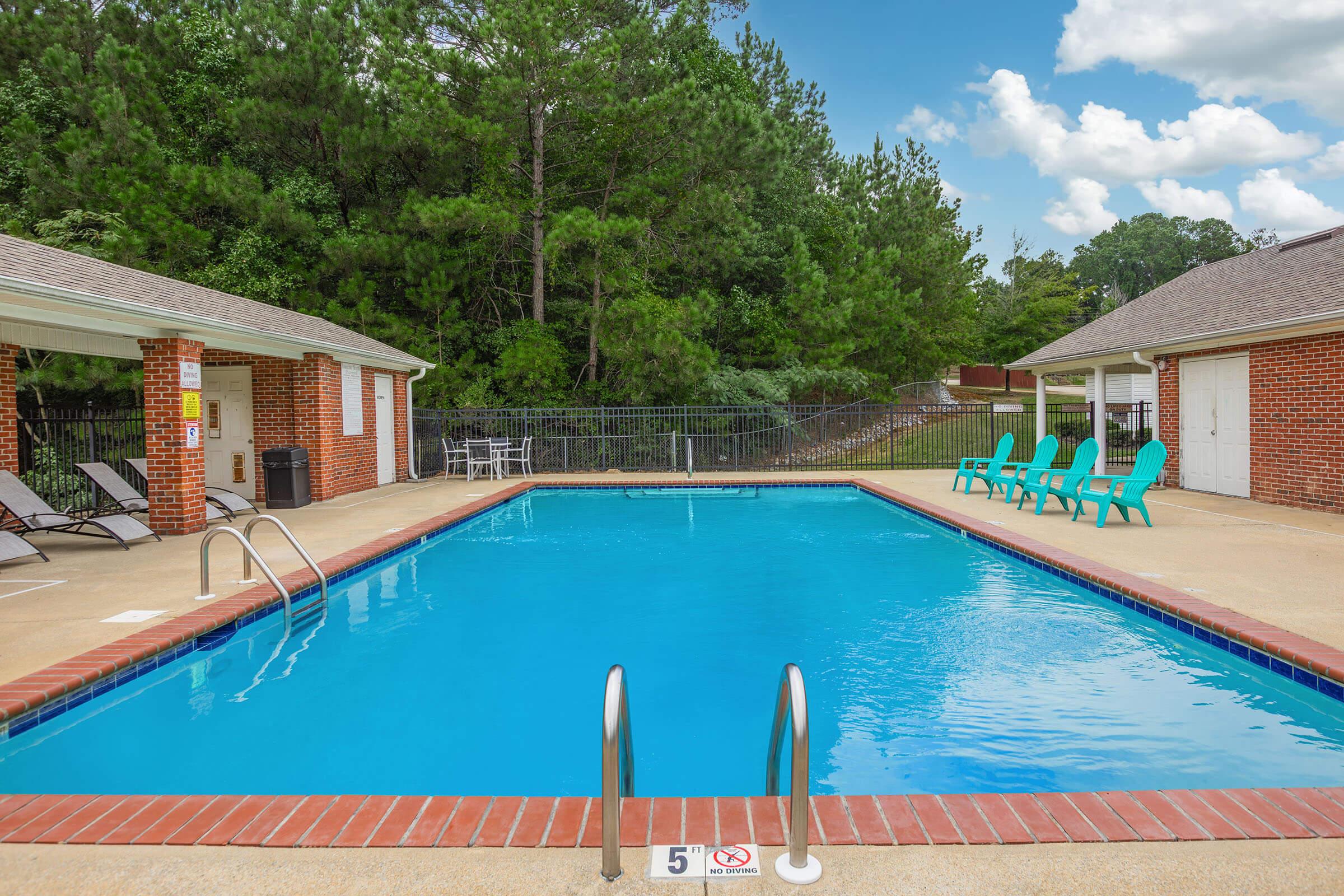 A clear swimming pool surrounded by a concrete deck, featuring lounge chairs and bright green poolside chairs. The backdrop includes trees and a blue sky with fluffy white clouds. Nearby, there are two buildings with brick exteriors. A safety sign is visible near the pool edge.