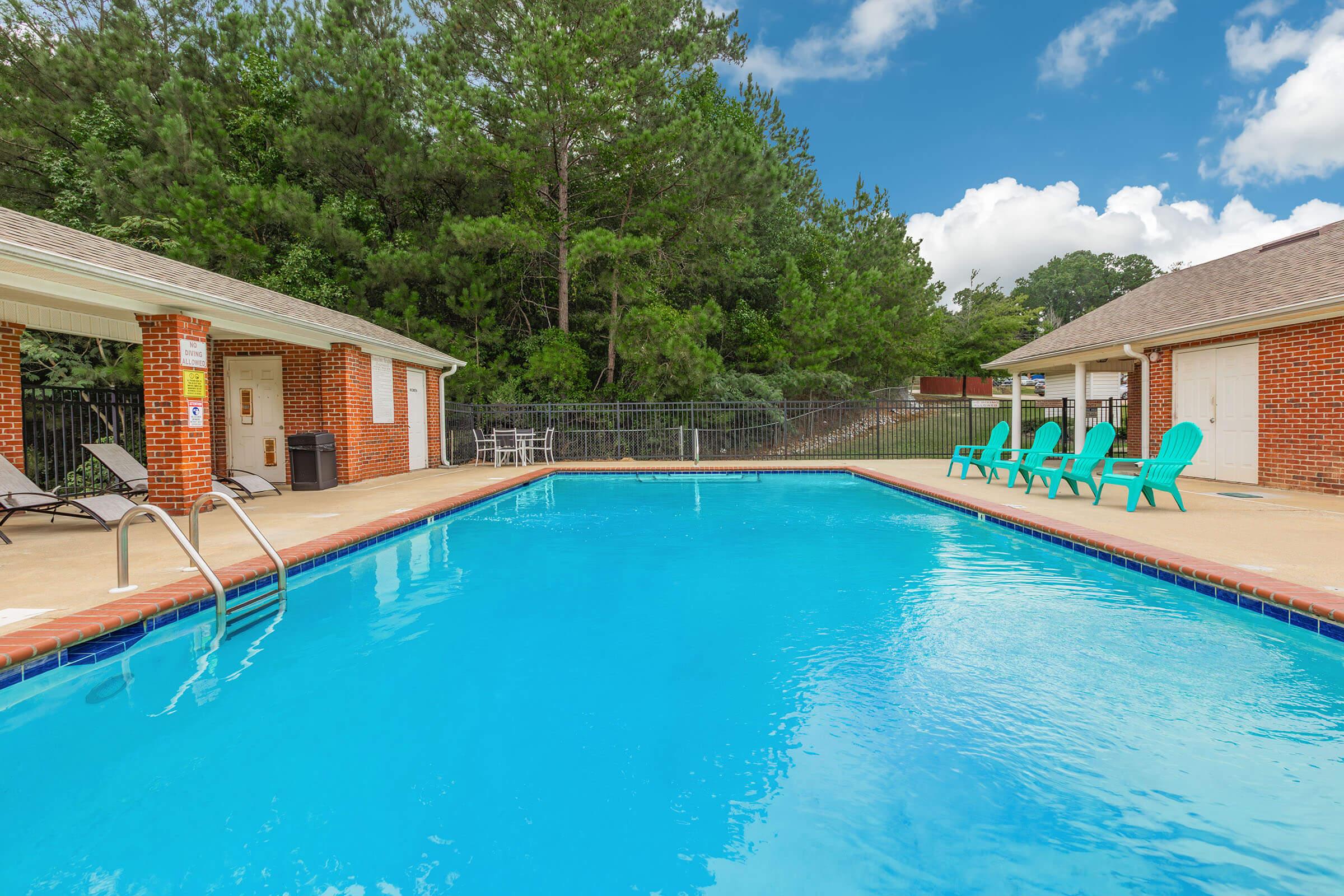 A clear blue swimming pool surrounded by a concrete deck. On one side, there are two brick buildings, each with a white door. Green lawn and trees are visible in the background. Several turquoise lounge chairs are positioned beside the pool. The sky is partly cloudy.