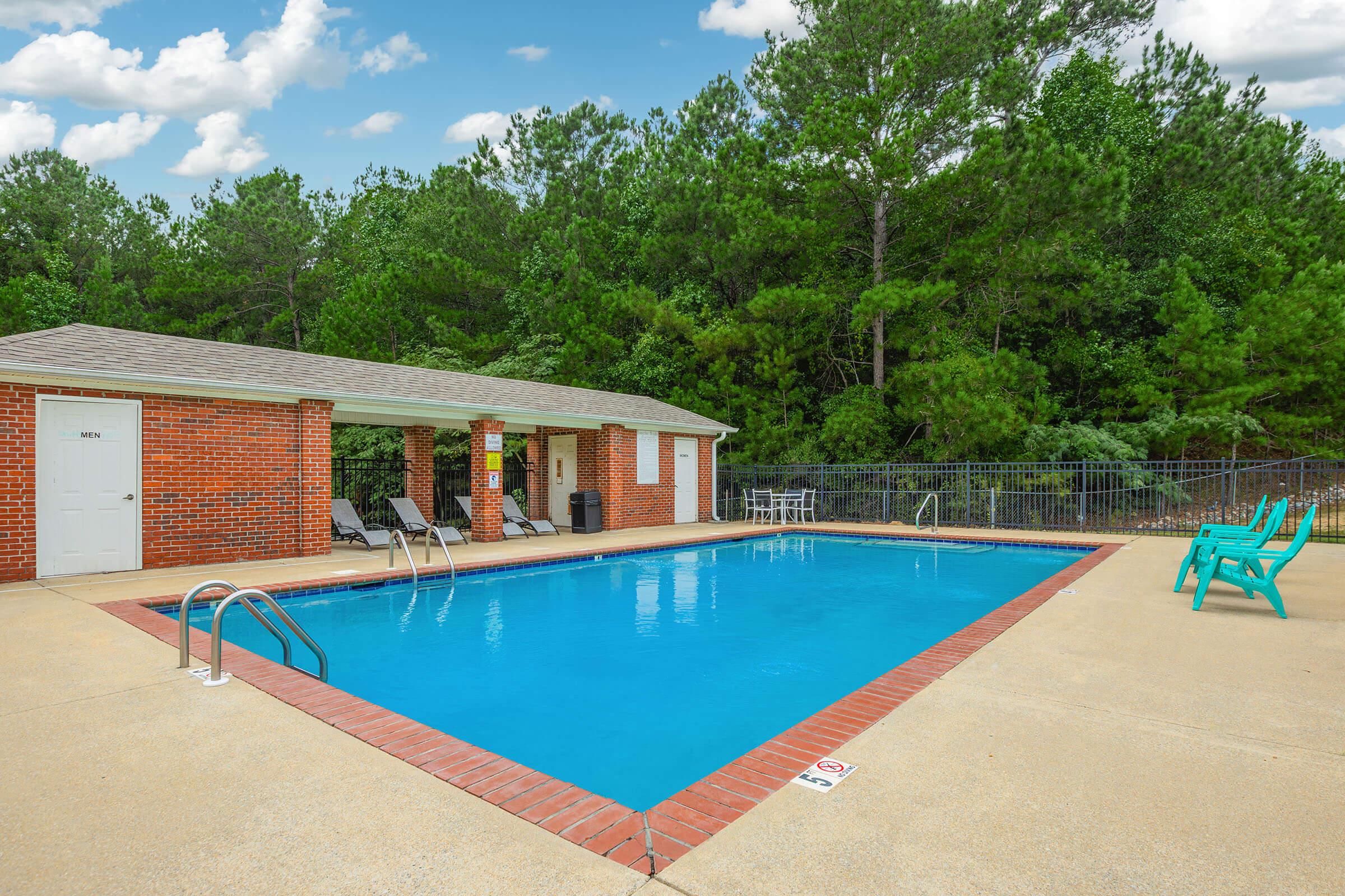A clear blue swimming pool surrounded by a concrete deck. There are several lounge chairs positioned around the pool, with a small shaded area featuring a roofed structure. Lush green trees are visible in the background, under a partly cloudy sky.