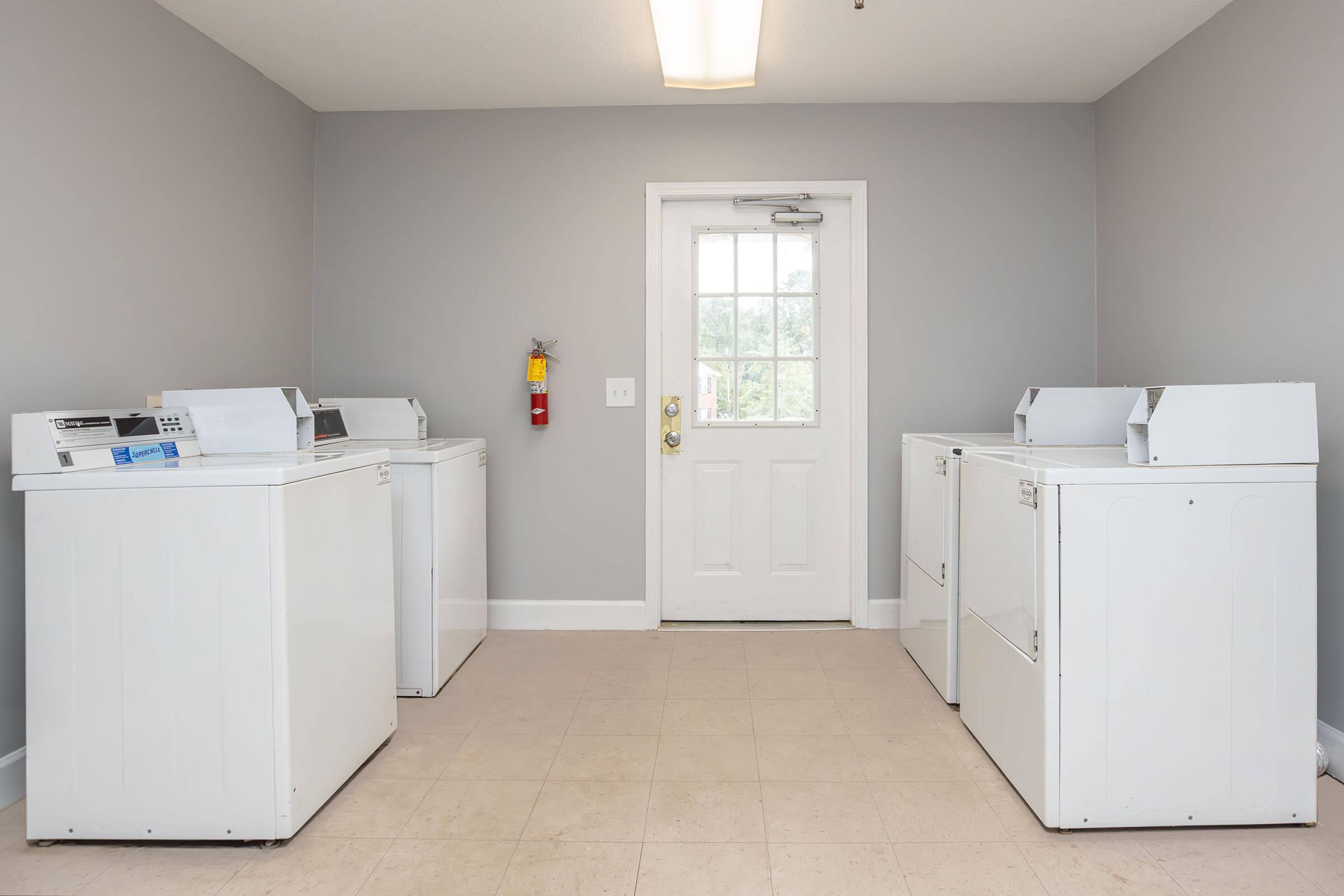 A clean laundry room featuring four white washing machines and a door with a window. The walls are painted gray and the floor is tiled. A fire extinguisher is mounted on the wall near the door. Natural light enters through the door.