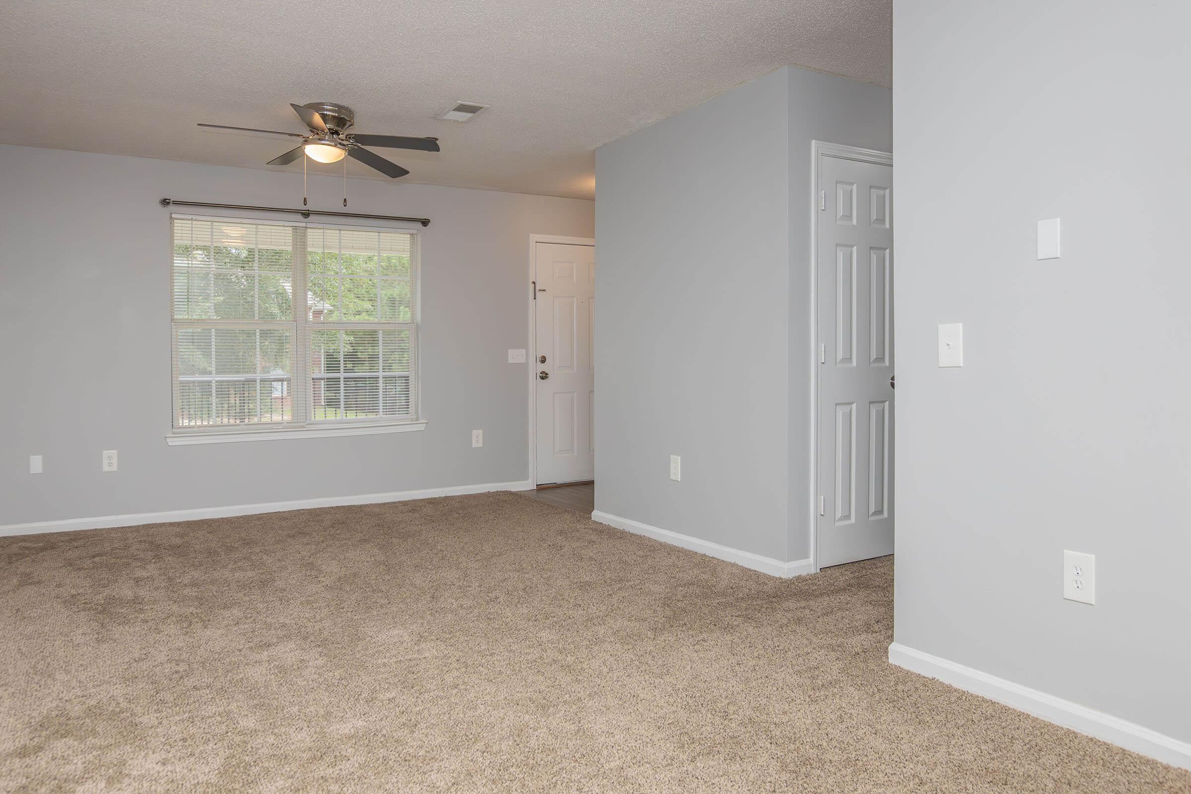 A spacious, carpeted living room featuring a ceiling fan, a large window with blinds, and light gray walls. A door leads outside, and there is a closed closet visible on the right. The room is well-lit and uncluttered, creating a welcoming atmosphere.
