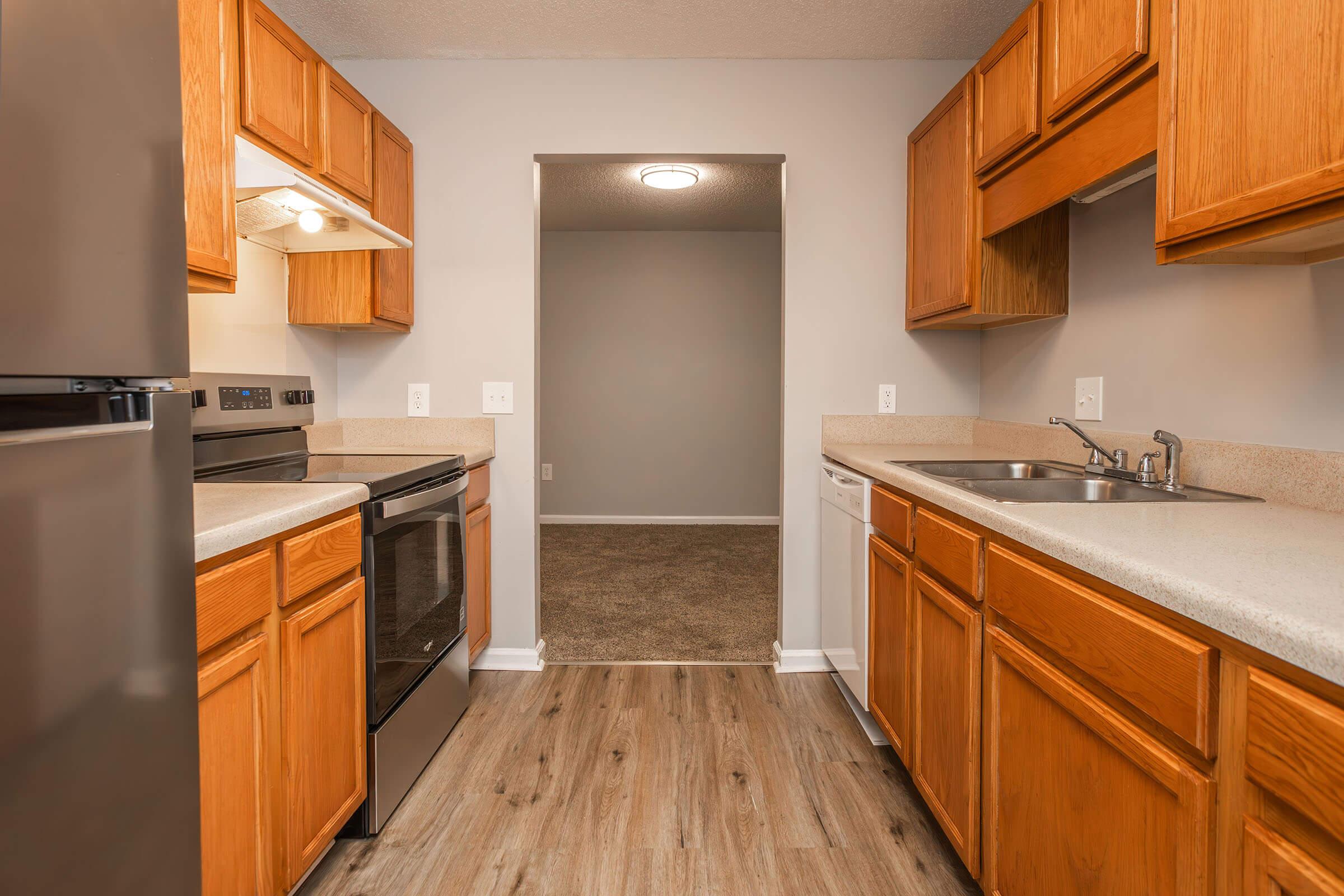 A modern kitchen featuring wooden cabinets, stainless steel appliances, and a light countertop. The space includes an oven, stovetop, microwave, and dishwasher, with a sink near the window. The wall is painted in a neutral color, and a doorway leads to a carpeted area beyond.