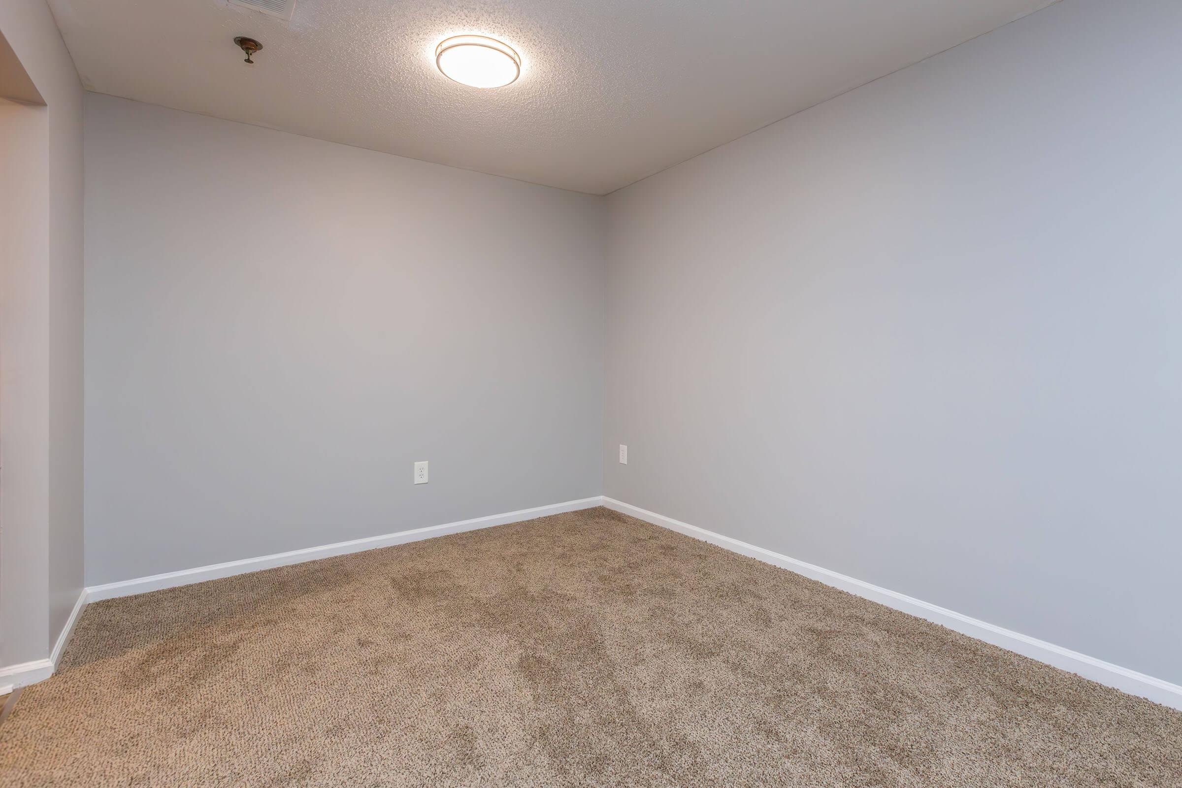 A vacant room with light gray walls and a carpeted floor. The ceiling features a round light fixture, and there are no furnishings or decor present. The space appears clean and minimalist, suggesting potential for various uses.