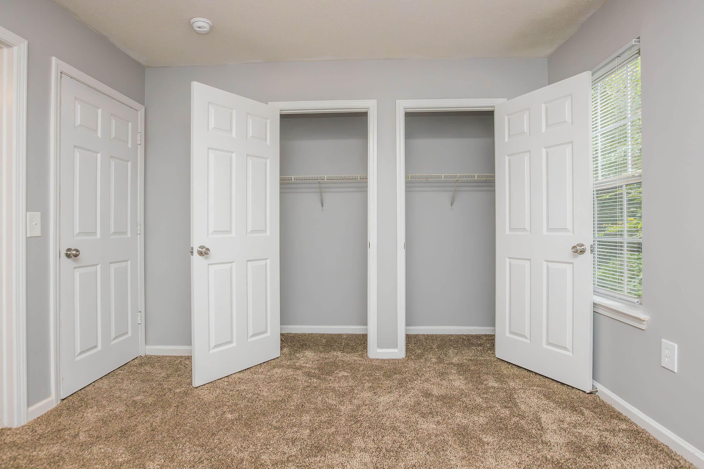 A spacious room featuring two open closet doors with hanging space, light gray walls, and windows allowing natural light. The floor is covered in plush beige carpet, creating a cozy and inviting atmosphere.