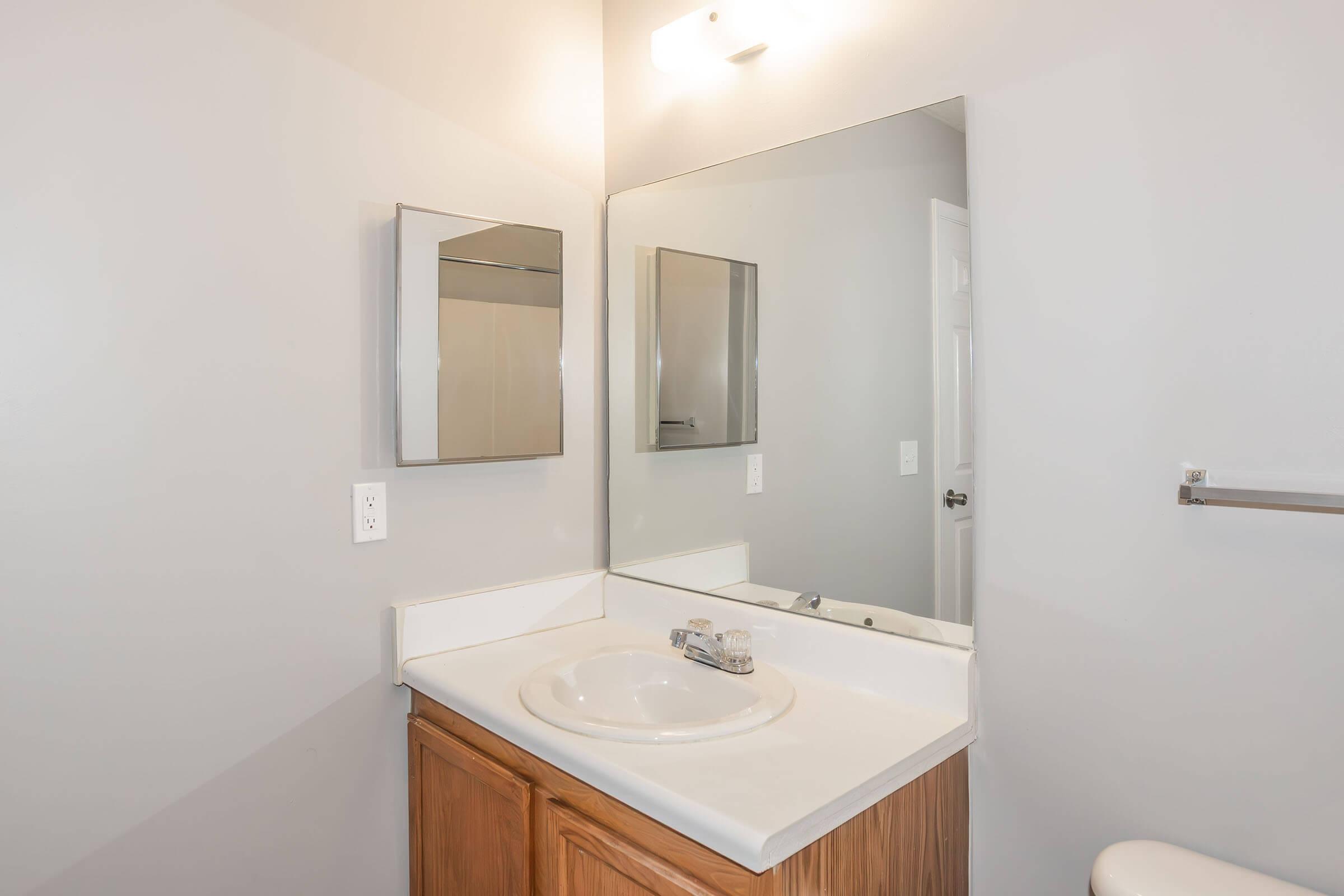 A modern bathroom with a light gray wall. It features a simple white countertop with a round sink, a mirror above the sink, and wooden cabinetry underneath. There is a toilet in the corner and a towel rack on the wall, creating a clean and functional design.
