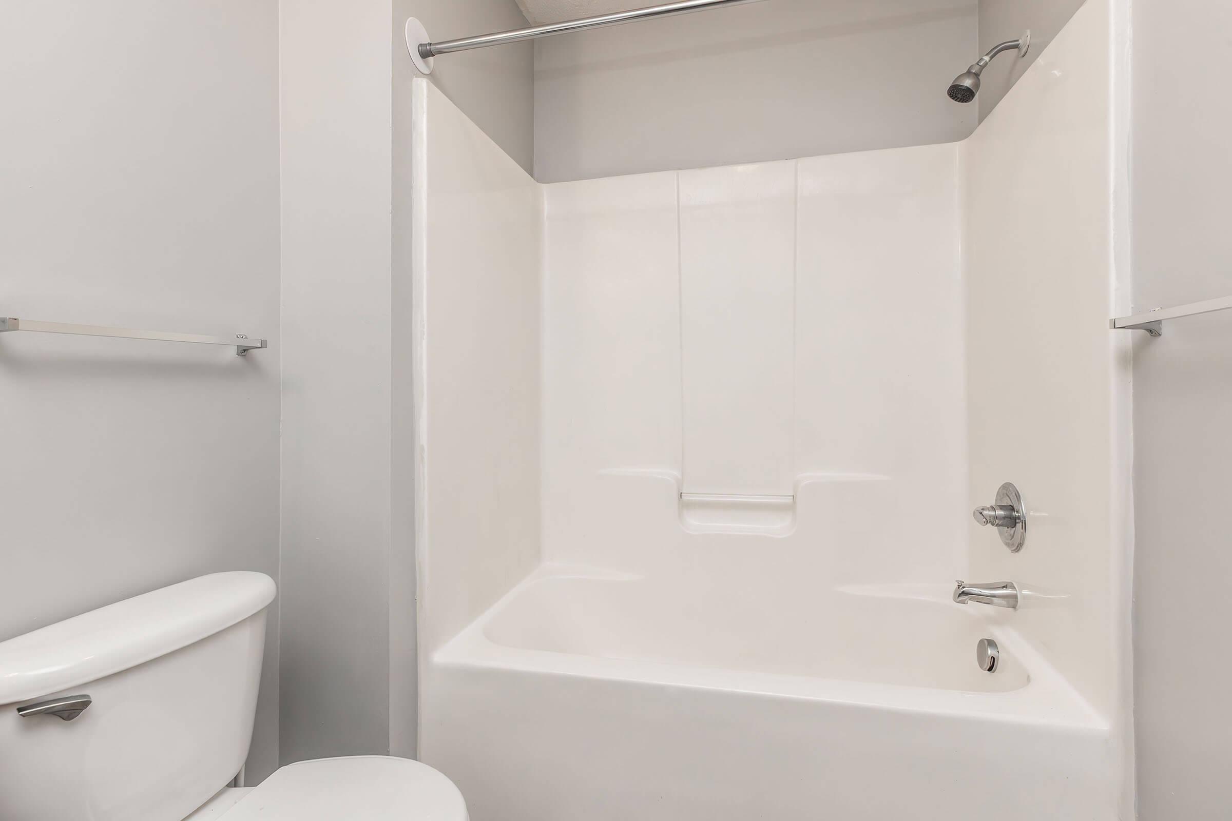 A clean, modern bathroom featuring a white bathtub and shower with a sleek chrome faucet, alongside a white toilet. The walls are painted light gray, and there are grab bars for safety. The overall appearance is bright and spacious, emphasizing simplicity and functionality.
