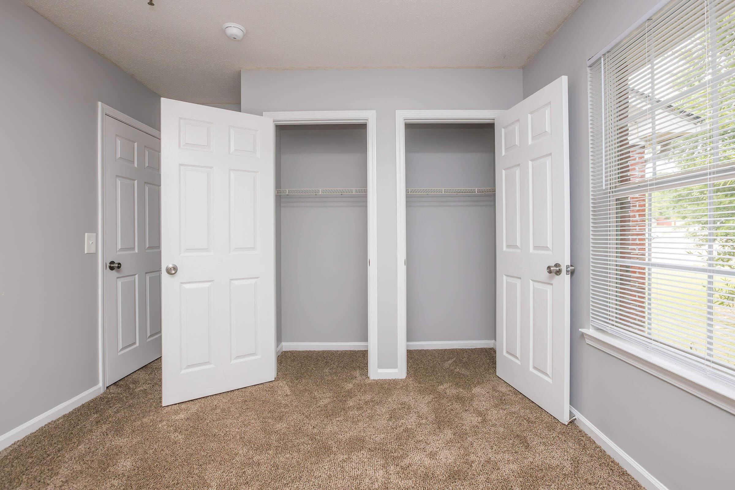 A well-lit room with two open closet doors revealing empty closets. The walls are painted light gray, and the floor is covered with carpet. A window with blinds allows natural light to enter, and the overall space appears clean and organized.
