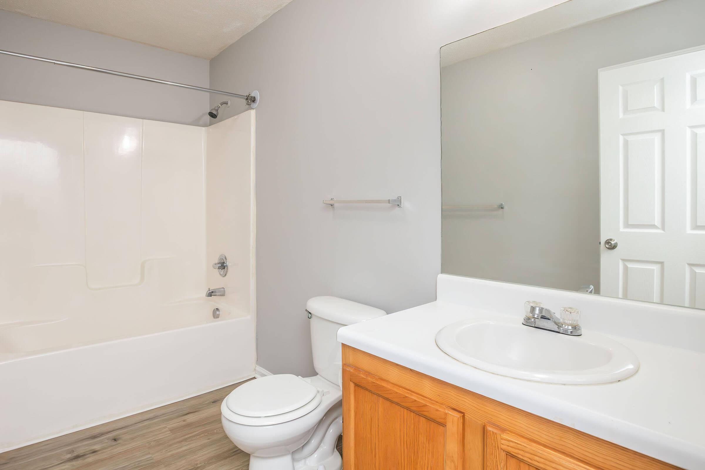 A clean and modern bathroom featuring a white bathtub with a shower rod, a toilet, and a sink with a wooden cabinet underneath. The walls are painted gray, and a large mirror is mounted above the sink. A white door is visible in the background, and the floor has a wooden appearance.