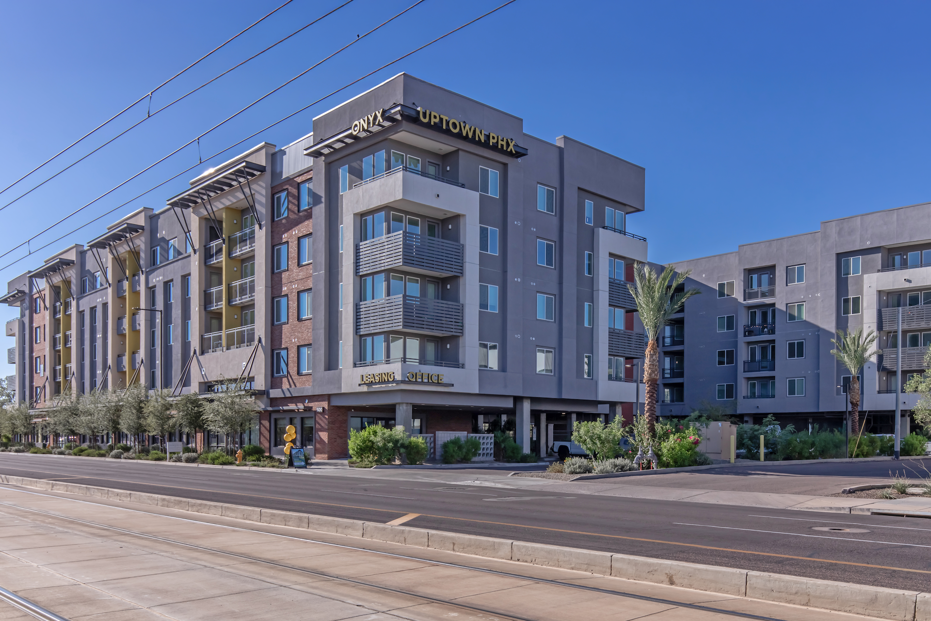 Modern residential building labeled "Uptown Phx" with multiple floors, large windows, and a mix of gray and red brick exteriors. Surrounding greenery and palm trees, with a clear blue sky overhead. Street with a sidewalk runs in front of the building.