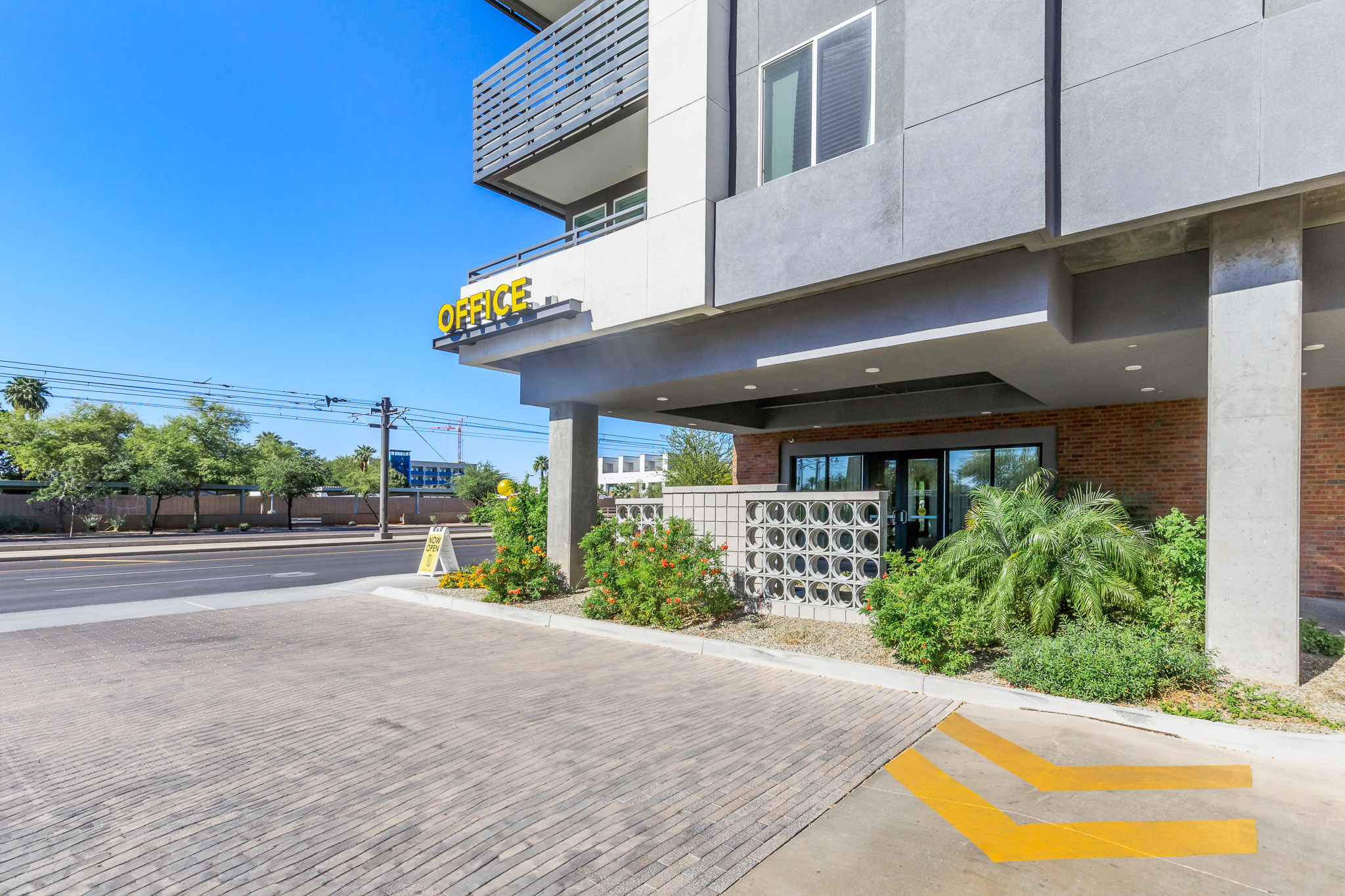 Modern office building exterior with large glass doors and a prominent "OFFICE" sign. The façade features a mix of concrete and brick materials. Lush greenery and decorative plants are arranged near the entrance, while a patterned concrete pathway leads to the building, with a clear blue sky above.