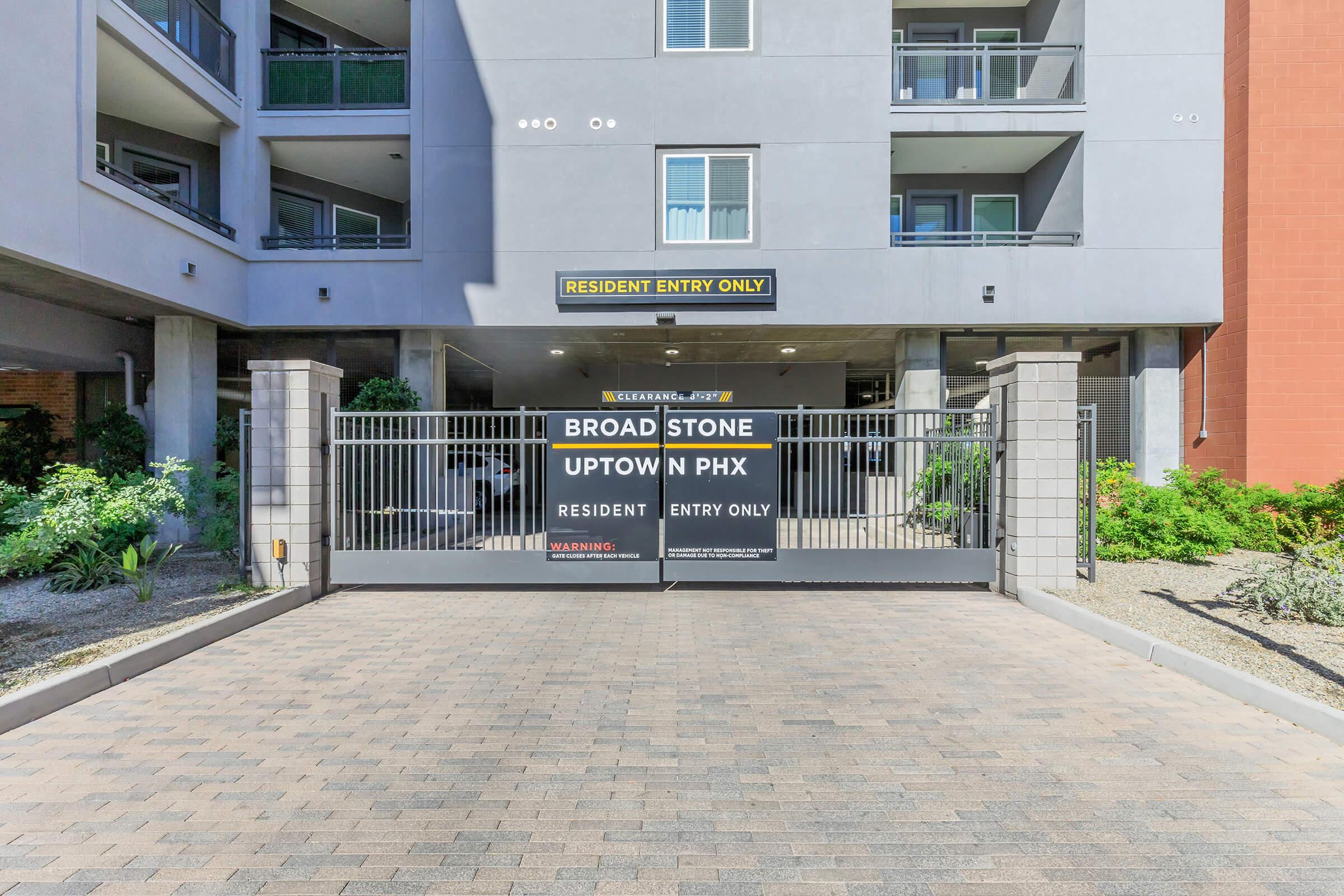 A gated entrance to Broad Stone Uptown in Phoenix, featuring a sign that reads "RESIDENT ENTRY ONLY" and another sign with the property name. The area is landscaped with plants, and there are residential buildings in the background. The ground is paved with stone.