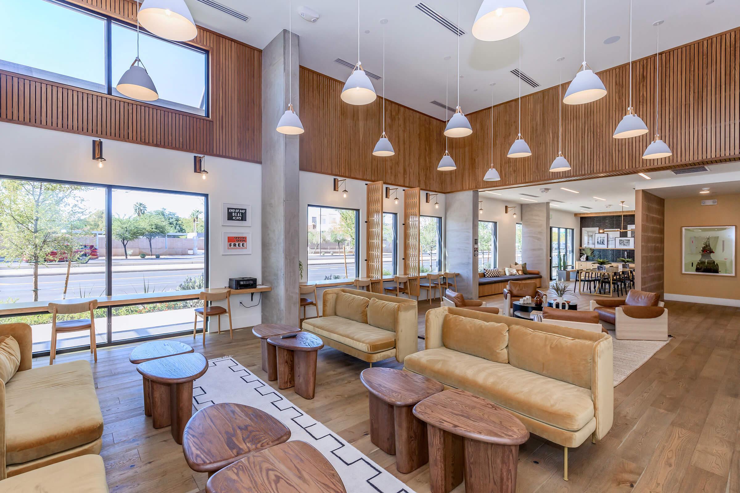 Interior of a modern lounge featuring large windows, wooden walls, and a mix of comfortable seating, including beige couches and round wooden tables. The space is bright and airy, with pendant lighting and a cozy atmosphere, designed for relaxation and socializing.