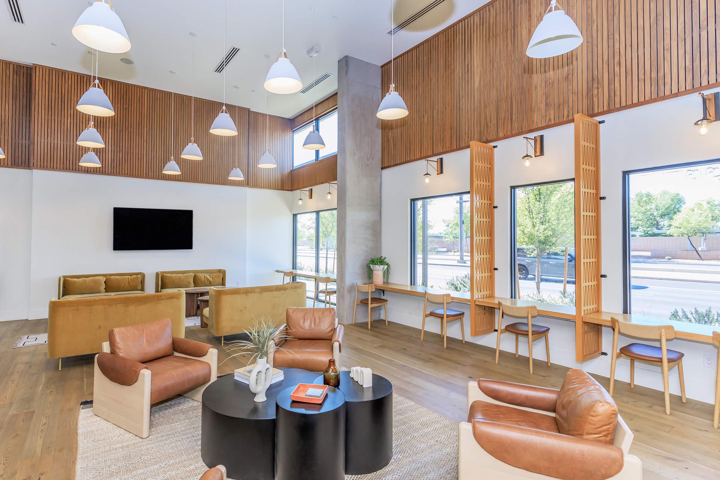 A modern lounge area featuring comfortable seating with tan leather chairs and a brown sofa. The space has large windows allowing natural light, wooden panel accents, and stylish pendant lights hanging from the ceiling. A round coffee table sits in the center, complemented by decorative elements.