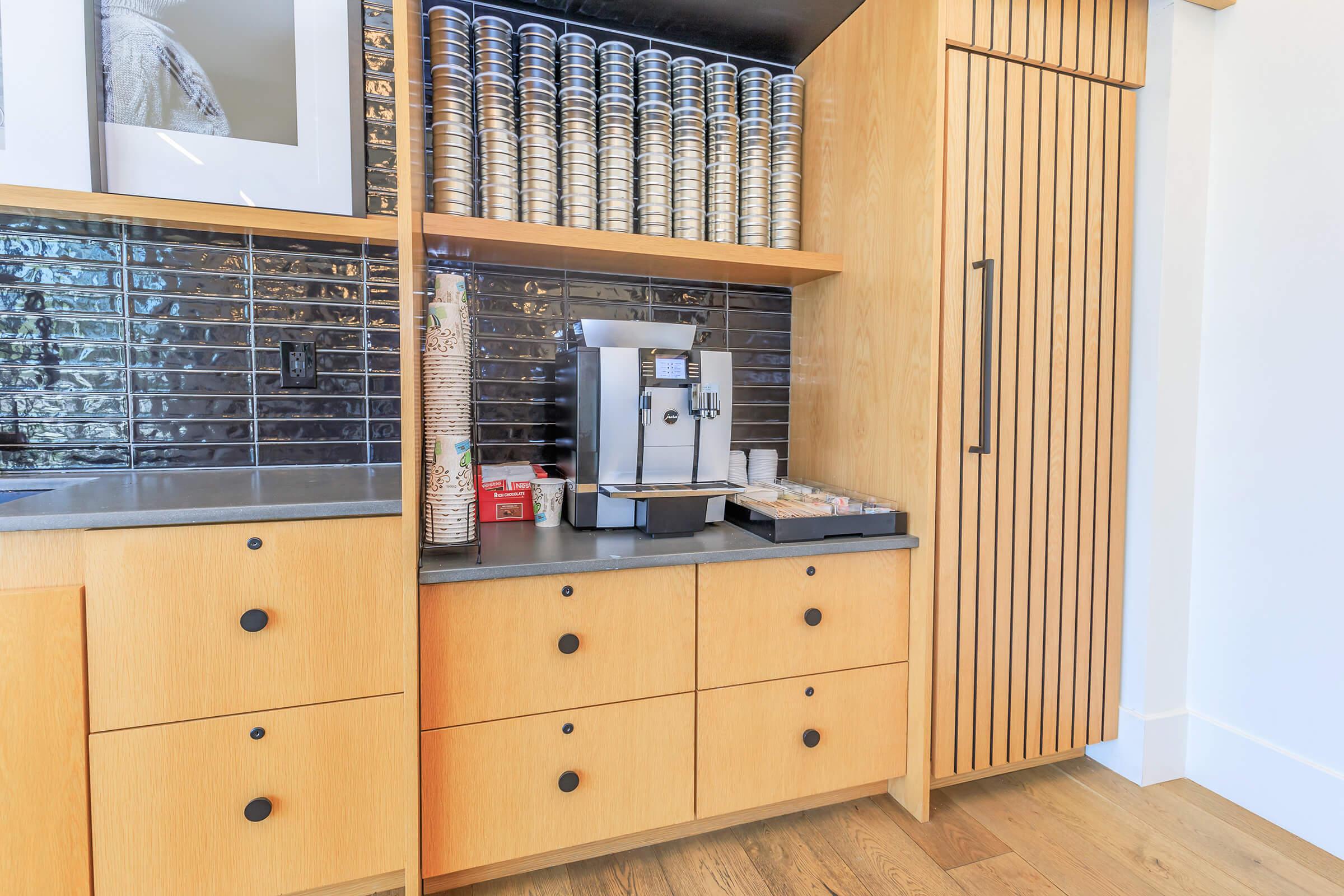 A modern kitchen setup featuring a wooden cabinetry with a coffee machine on the countertop, neatly arranged cups in a stack, and drawers below. The backsplash consists of sleek, dark tiles, adding elegance to the overall design. Natural light enhances the warm tones of the wood and flooring.