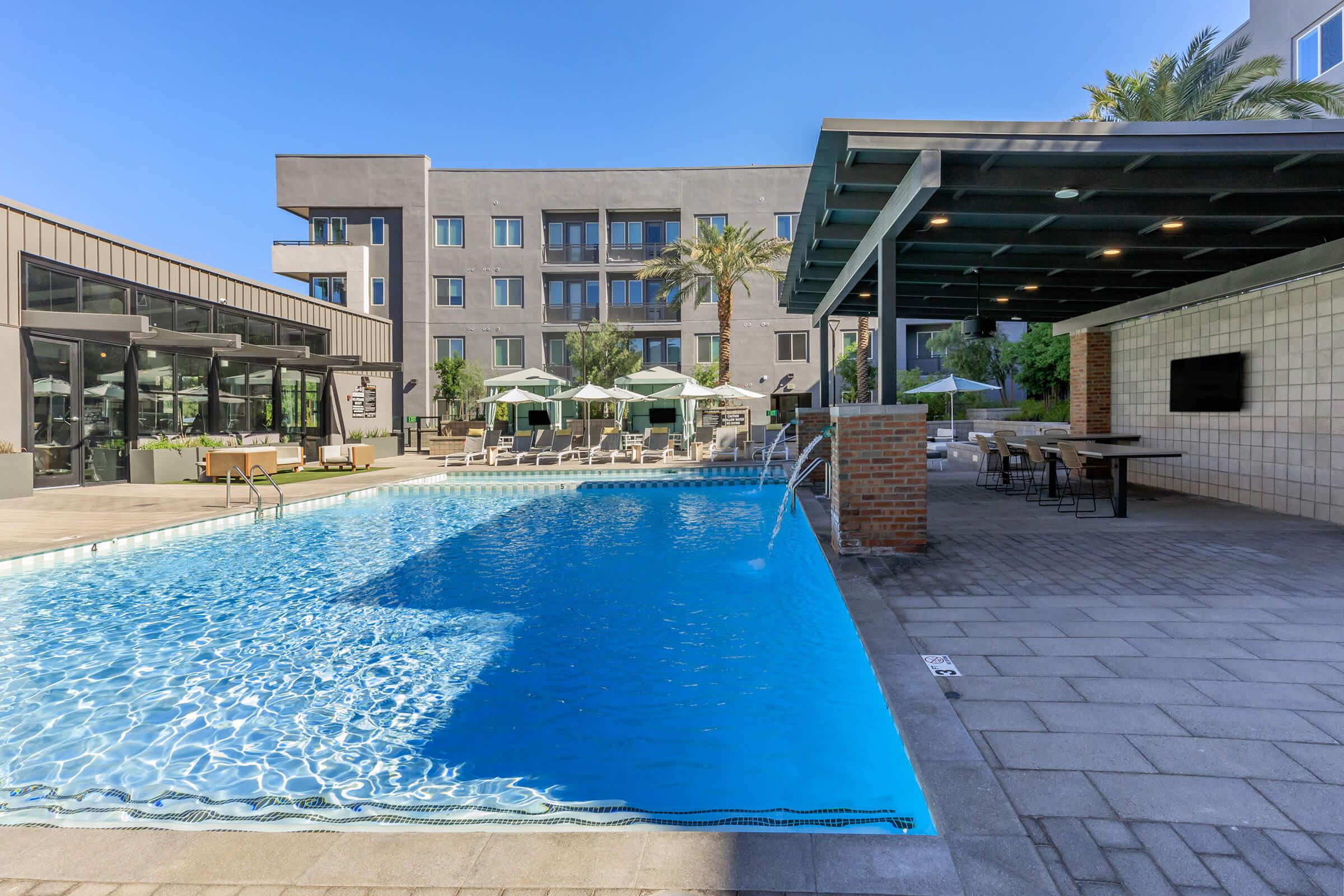 A serene swimming pool with clear blue water, surrounded by a spacious patio area. There are lounge chairs and shaded seating under umbrellas, along with modern building architecture in the background and palm trees adding to the tropical ambiance. Ideal for relaxation and leisure.
