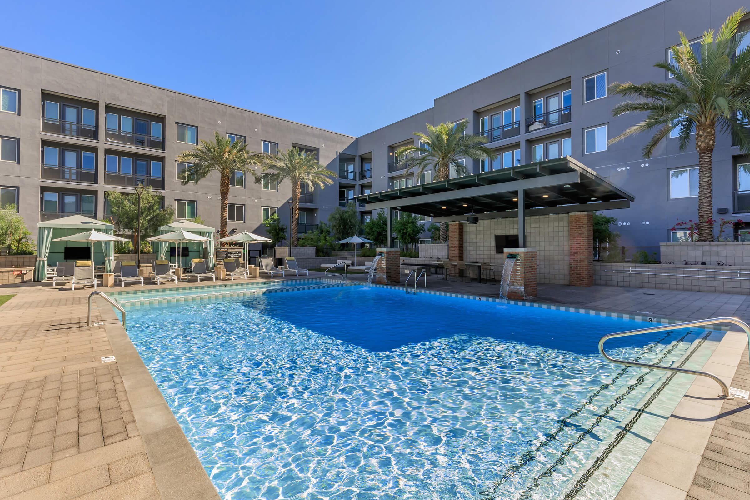 A spacious outdoor swimming pool surrounded by lounge chairs and umbrellas, set in a landscaped area with palm trees. The pool reflects the blue sky and adjacent modern apartment buildings, providing a serene atmosphere for relaxation and leisure.