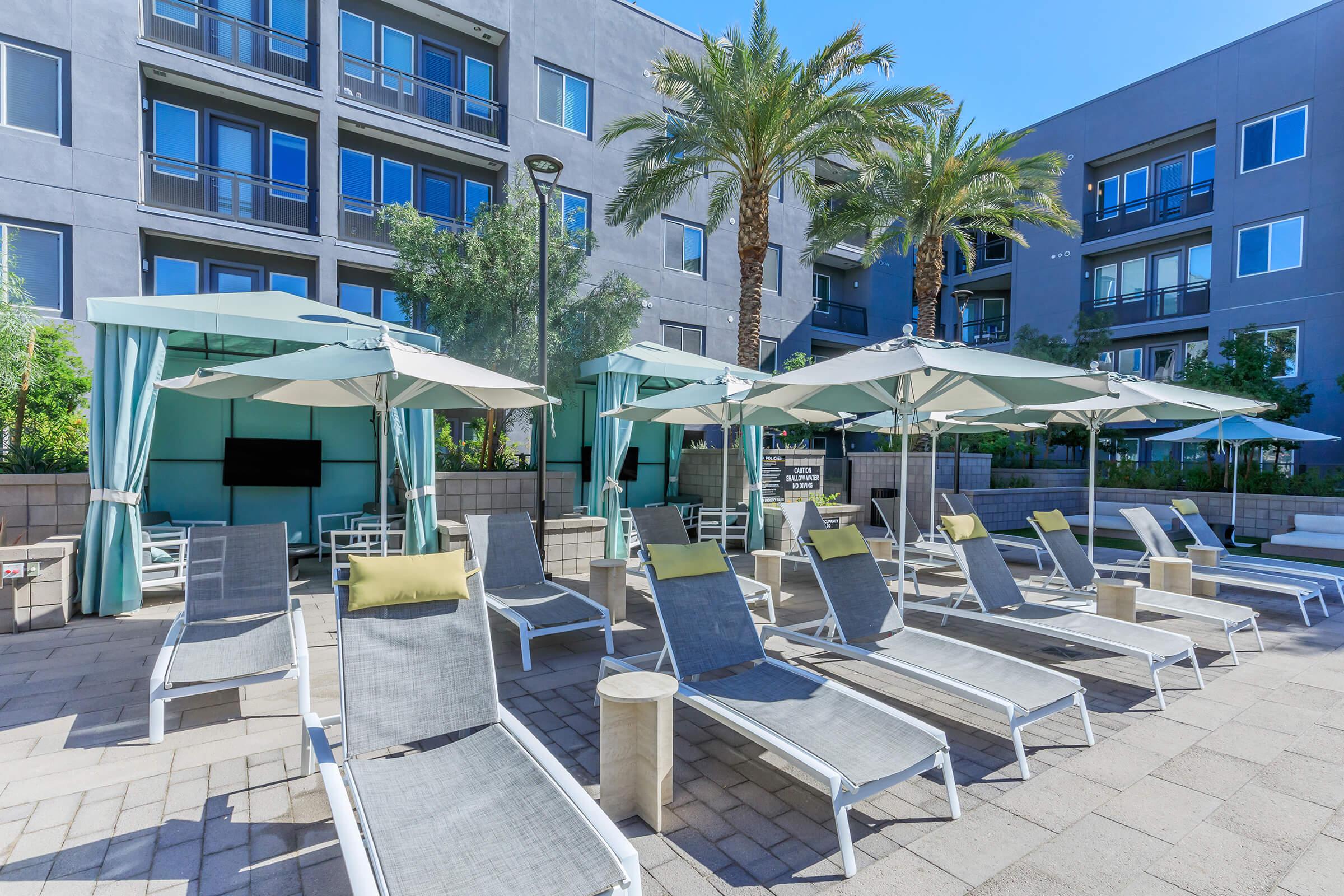 A resort-style pool area featuring numerous lounge chairs and umbrellas, surrounded by palm trees and modern apartment buildings. The space includes cabanas and a flat-screen TV, creating a relaxed atmosphere for outdoor leisure. The layout showcases a sunny day with clear blue skies.