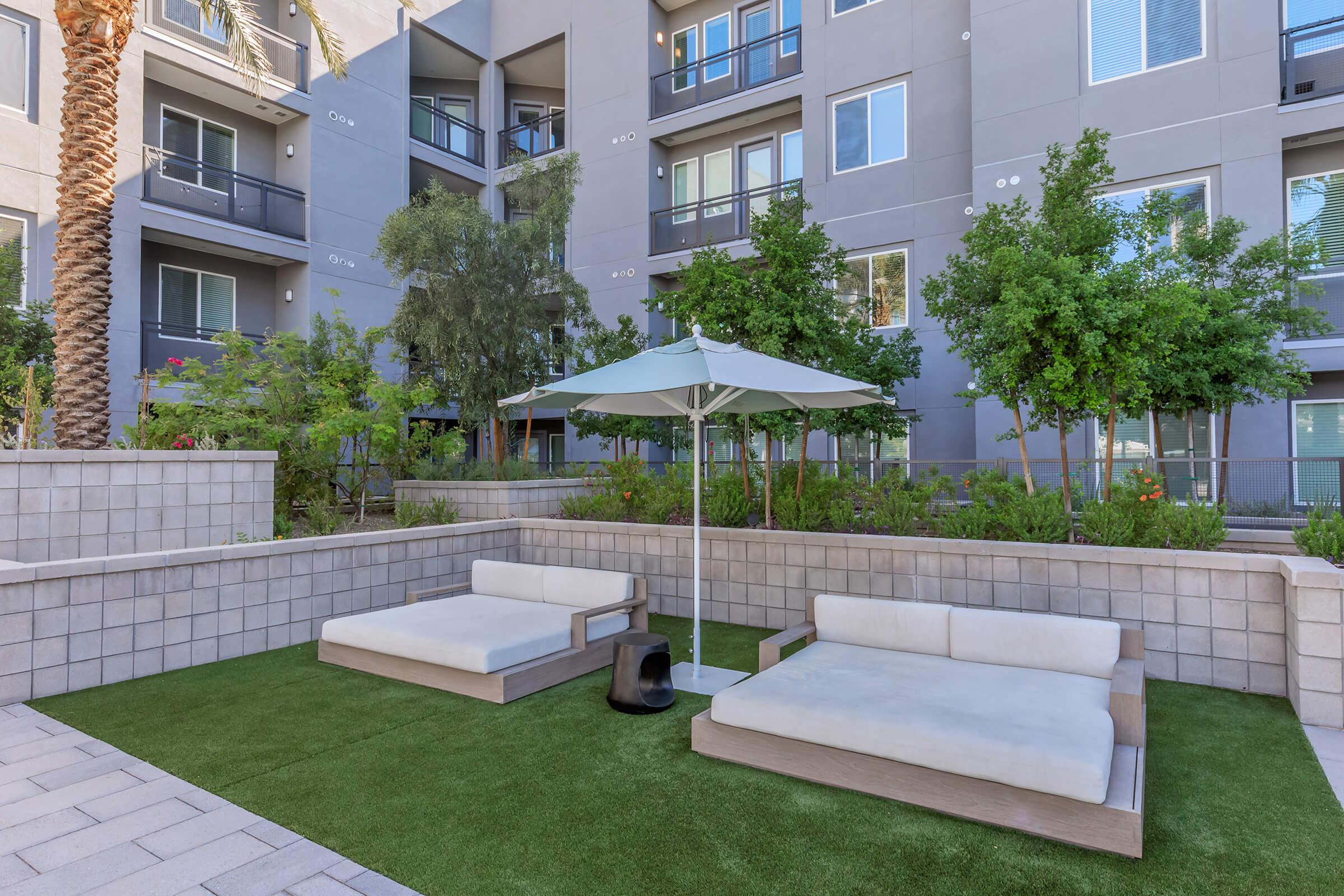 A peaceful outdoor lounge area featuring two padded chaise lounges under a white umbrella, surrounded by lush greenery and modern apartment buildings in the background. The space has a light-colored stone patio and artificial grass, creating a serene atmosphere for relaxation.