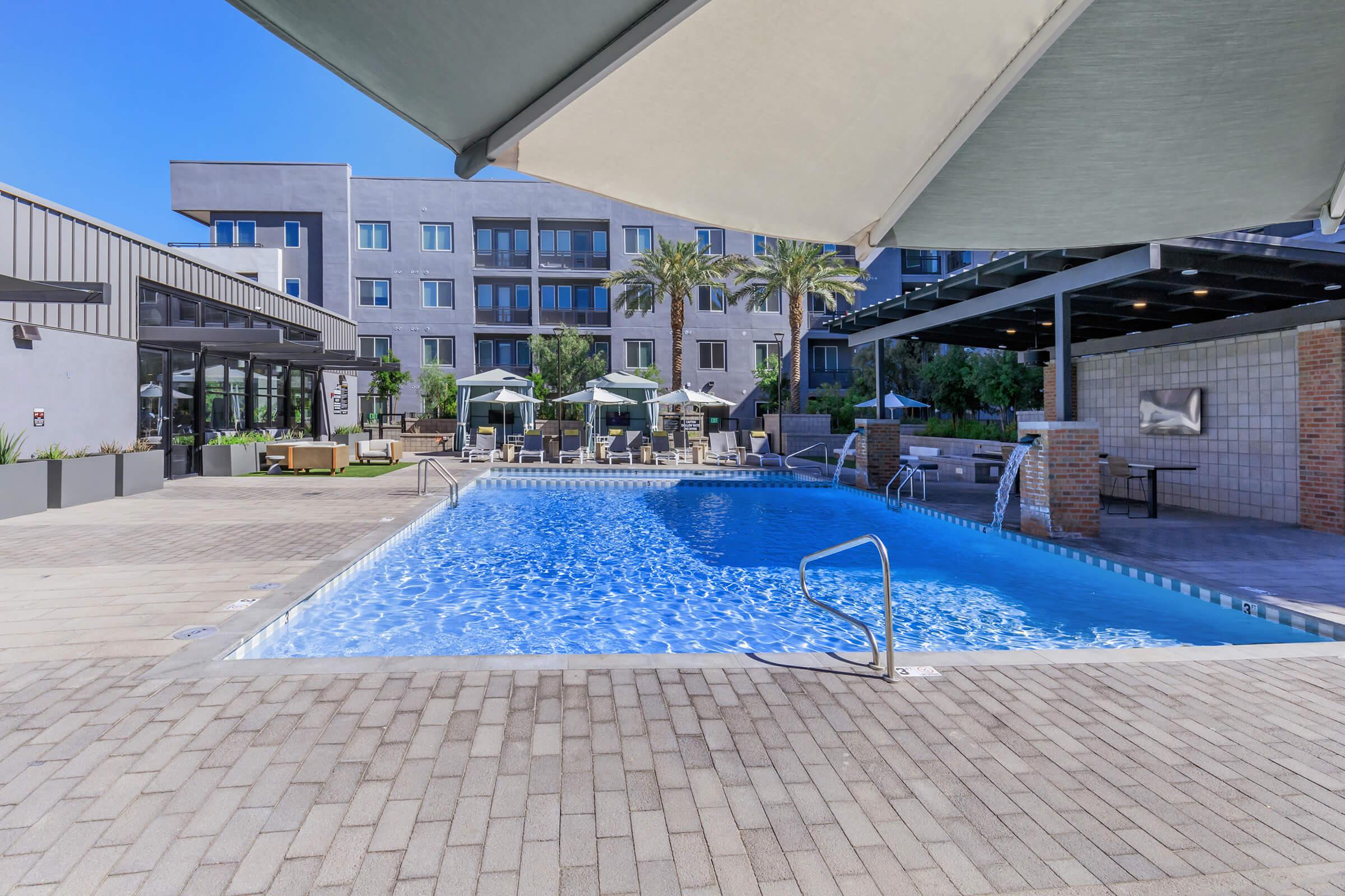 A bright and inviting outdoor pool area at a modern apartment complex, featuring a clear blue pool, comfortable lounge chairs, and shaded umbrellas. Surrounding the pool are lush palm trees and stylish seating areas, creating a relaxing atmosphere for residents and guests.