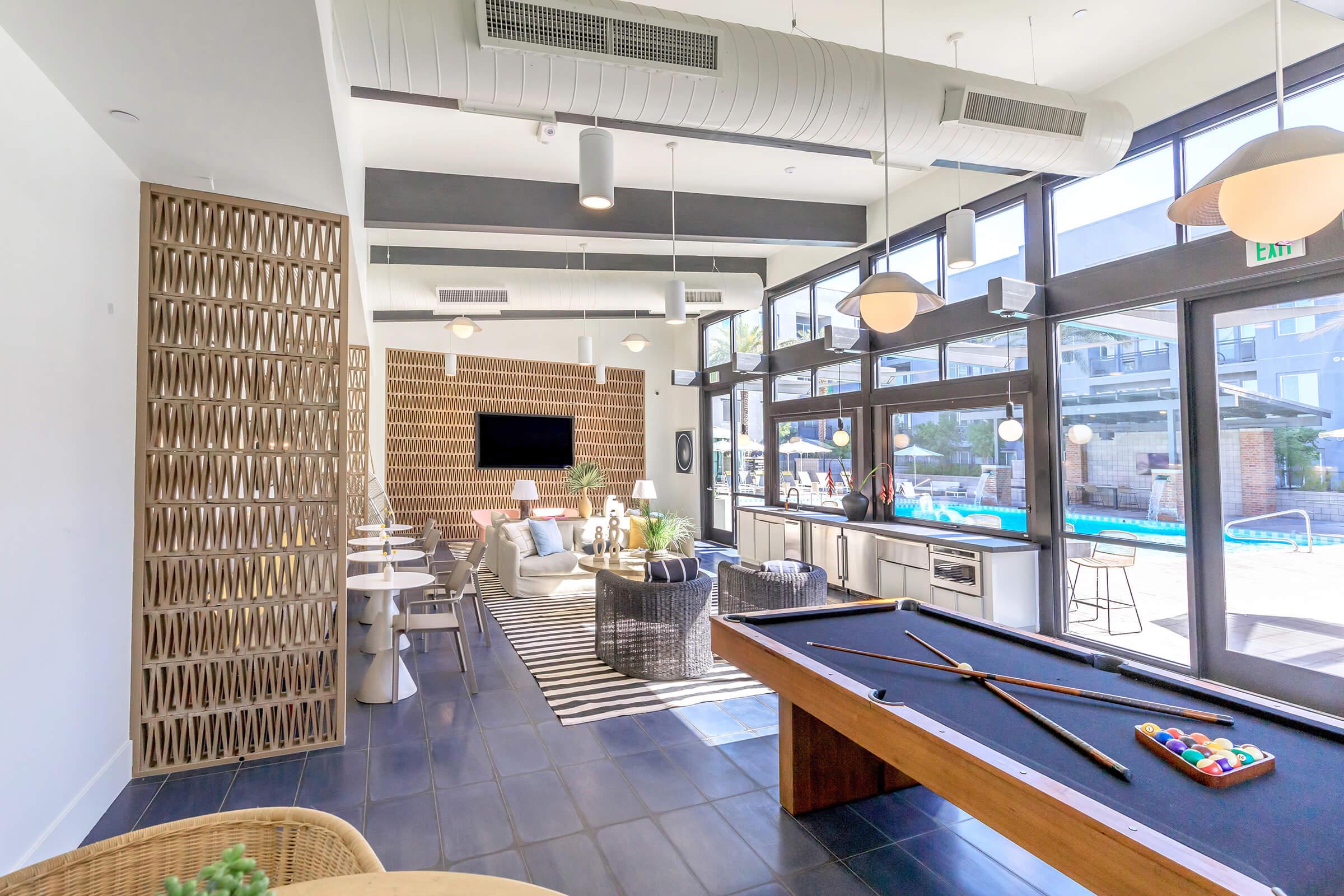 A spacious indoor lounge area featuring large windows with natural light, modern furniture, a pool table, and dining tables. The decor includes a stylish black and white rug, soft seating, and a wall-mounted TV, creating a relaxing environment that opens up to a poolside view.