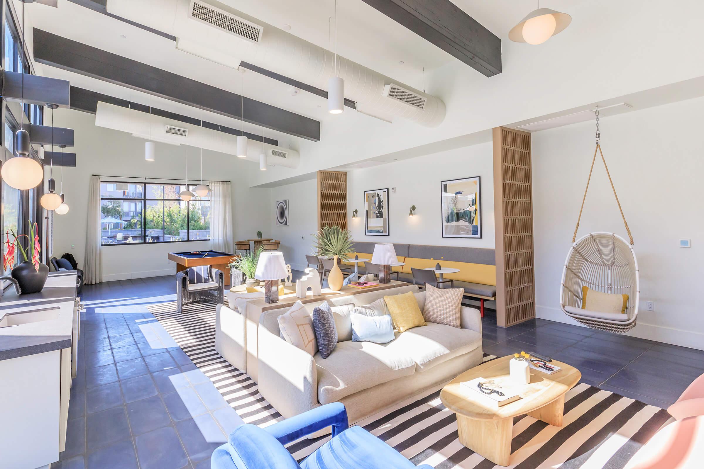 A bright and airy living space featuring a cozy sofa with decorative pillows, a wooden coffee table, and a hanging chair. Large windows let in natural light, illuminating the striped rug on the floor and modern decor elements. A dining area and pool table are visible in the background, enhancing the social atmosphere.
