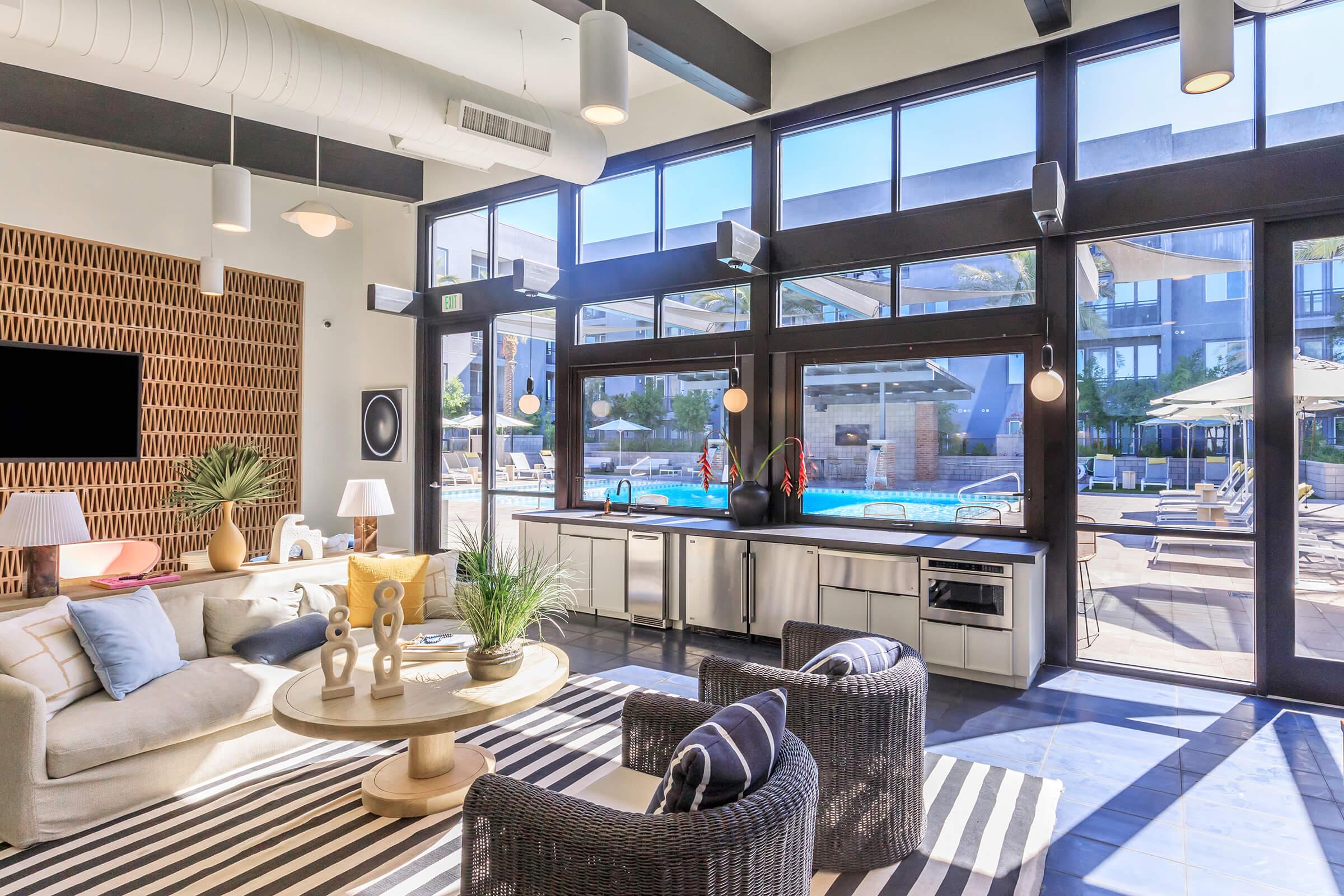 A spacious modern lounge area featuring a comfortable sofa, a round coffee table, and large windows that overlook a pool area. The decor includes indoor plants, stylish lighting, and a contemporary design with a striped rug, creating a bright and inviting atmosphere.