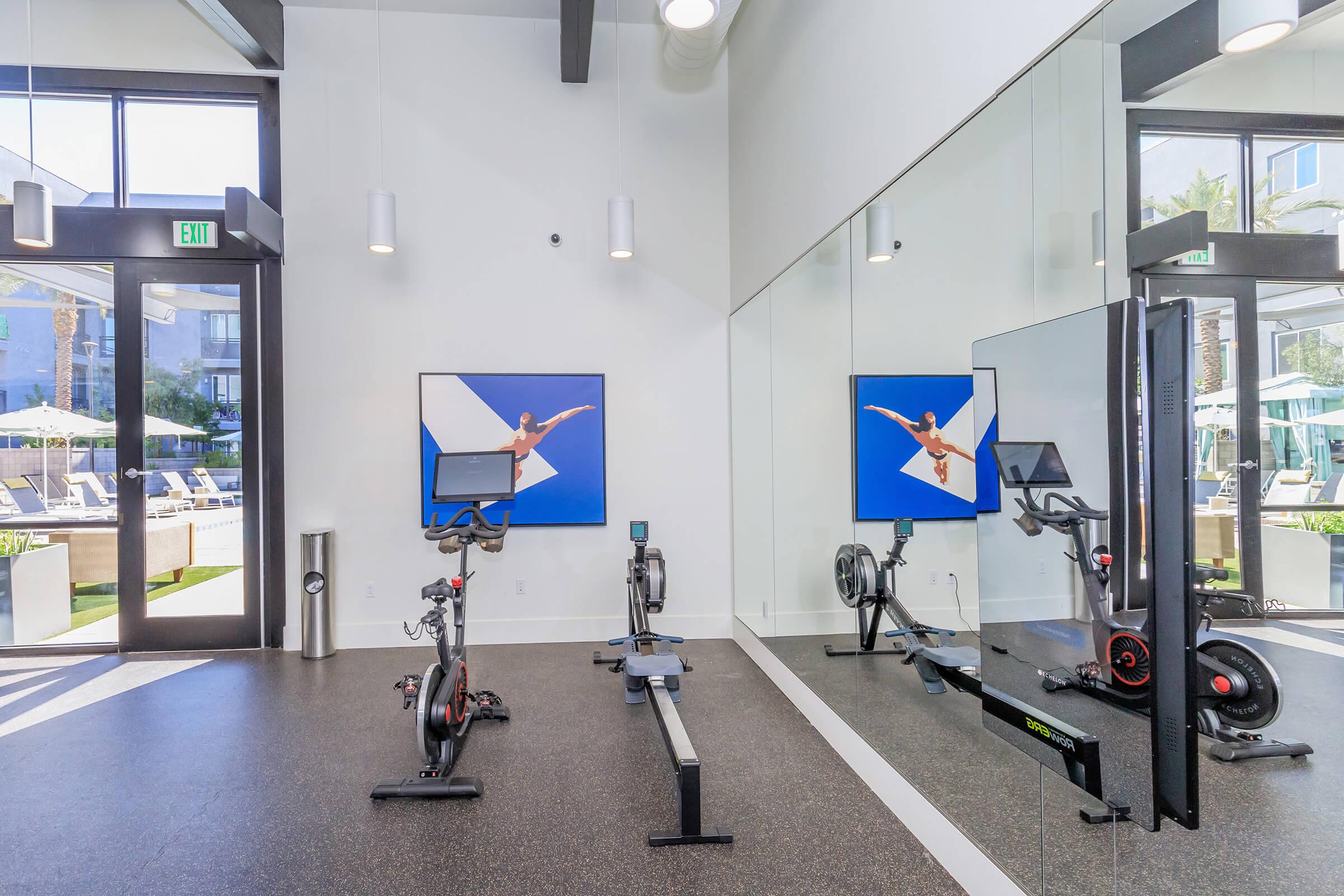 A modern gym interior featuring exercise equipment such as a stationary bike and rowing machine. Bright and spacious with large windows, the room includes a mirrored wall and is decorated with colorful artwork. The atmosphere is inviting, making it suitable for workouts.