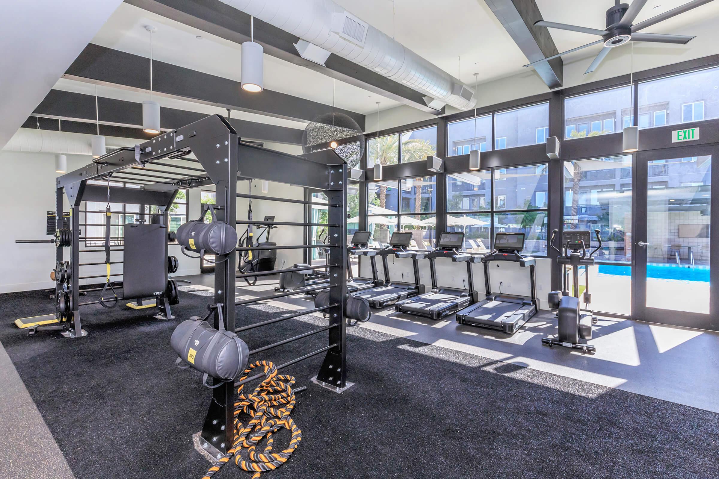 A modern gym featuring a variety of fitness equipment including treadmills, a weight training rig, and resistance bands. Large windows provide natural light, and the space has a sleek design with black flooring and ceiling fans, creating an inviting workout environment.