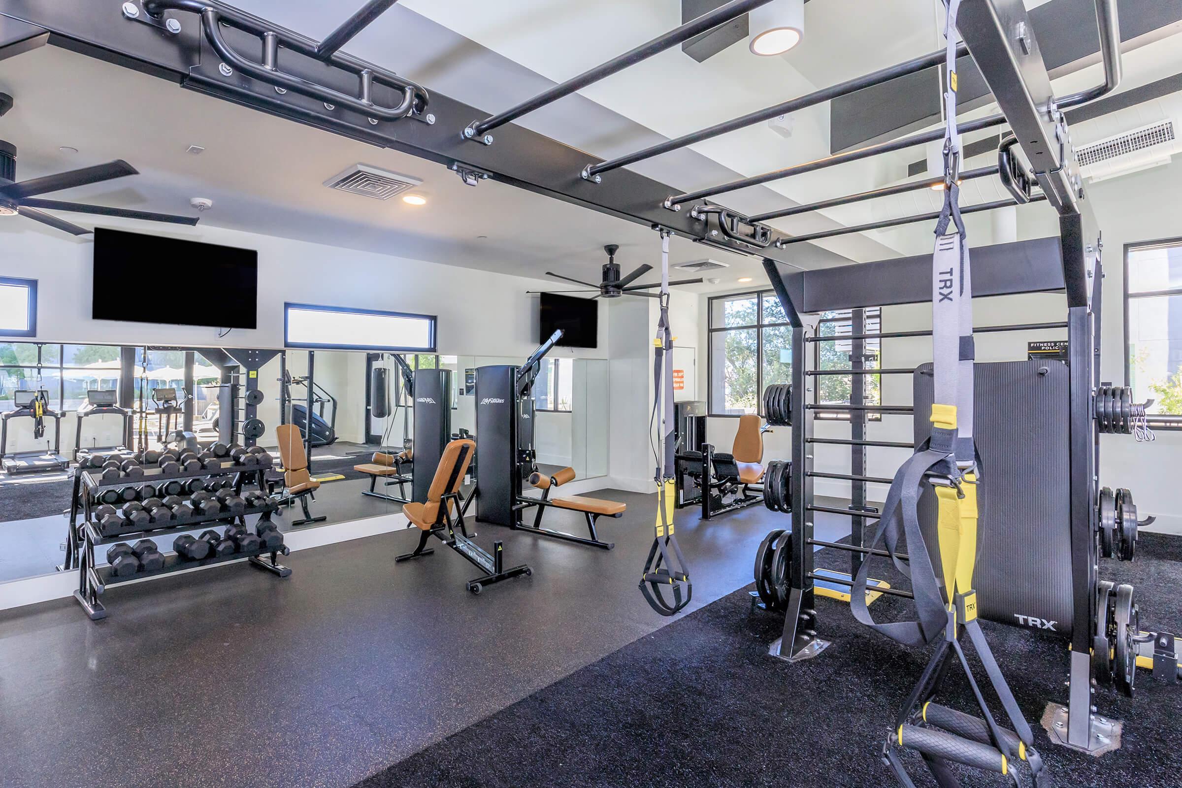 A modern fitness gym featuring weightlifting equipment, including dumbbells and a bench press, alongside a TRX suspension training system. The space has large windows for natural light, mirrors on the walls, and two flat-screen TVs mounted for workout guidance.