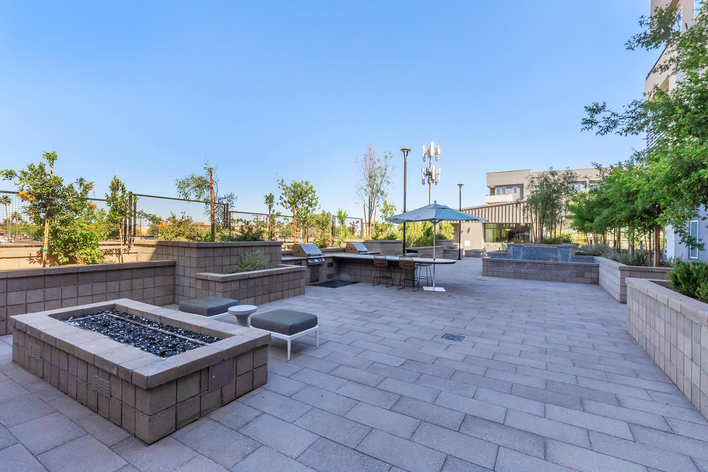 A spacious outdoor patio area featuring stone planters, lounge chairs, and a shaded umbrella. The environment is surrounded by greenery, with clear blue skies above, providing a relaxed and inviting atmosphere for socializing or enjoying leisure time.