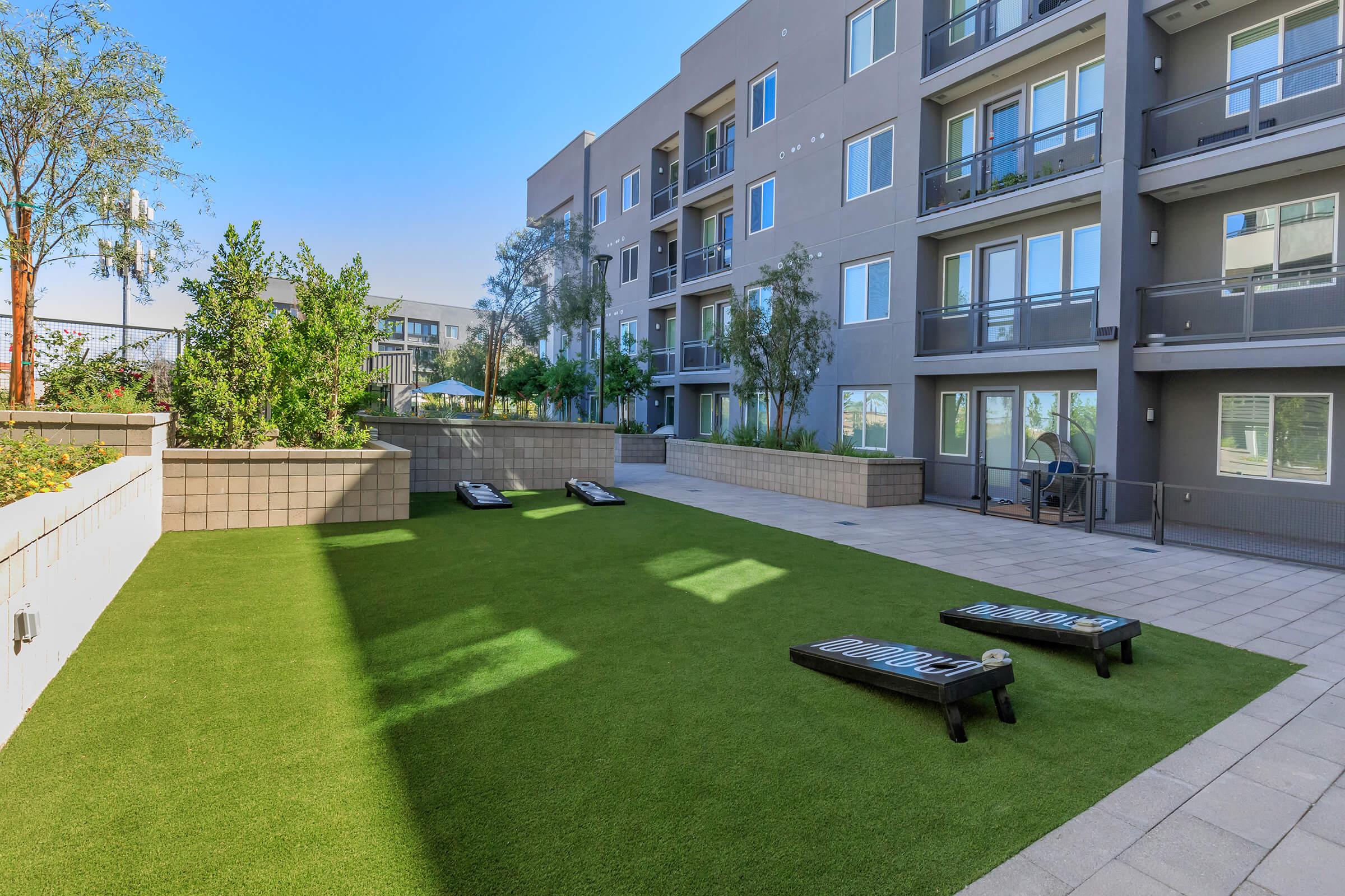 A landscaped outdoor area featuring artificial grass, flanked by modern apartment buildings. There are two lounge chairs on the grass, and surrounding plants and trees add greenery to the space. The sky is clear and blue, creating a bright, inviting atmosphere.