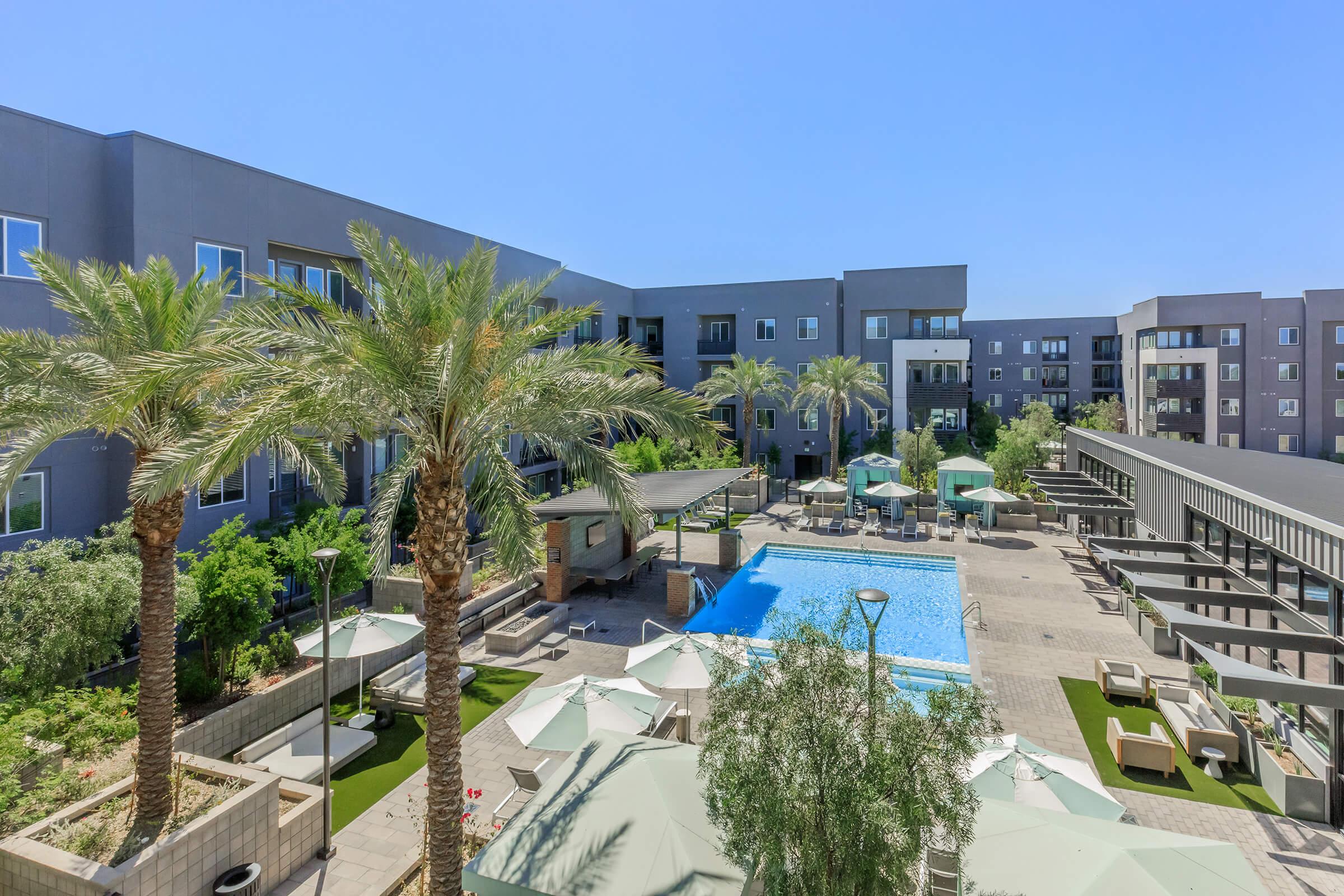 An expansive view of a hotel courtyard featuring a large swimming pool surrounded by palm trees, lounge chairs, and umbrellas. The modern architecture of the hotel buildings frames the scene under a clear blue sky, creating a relaxing ambiance.