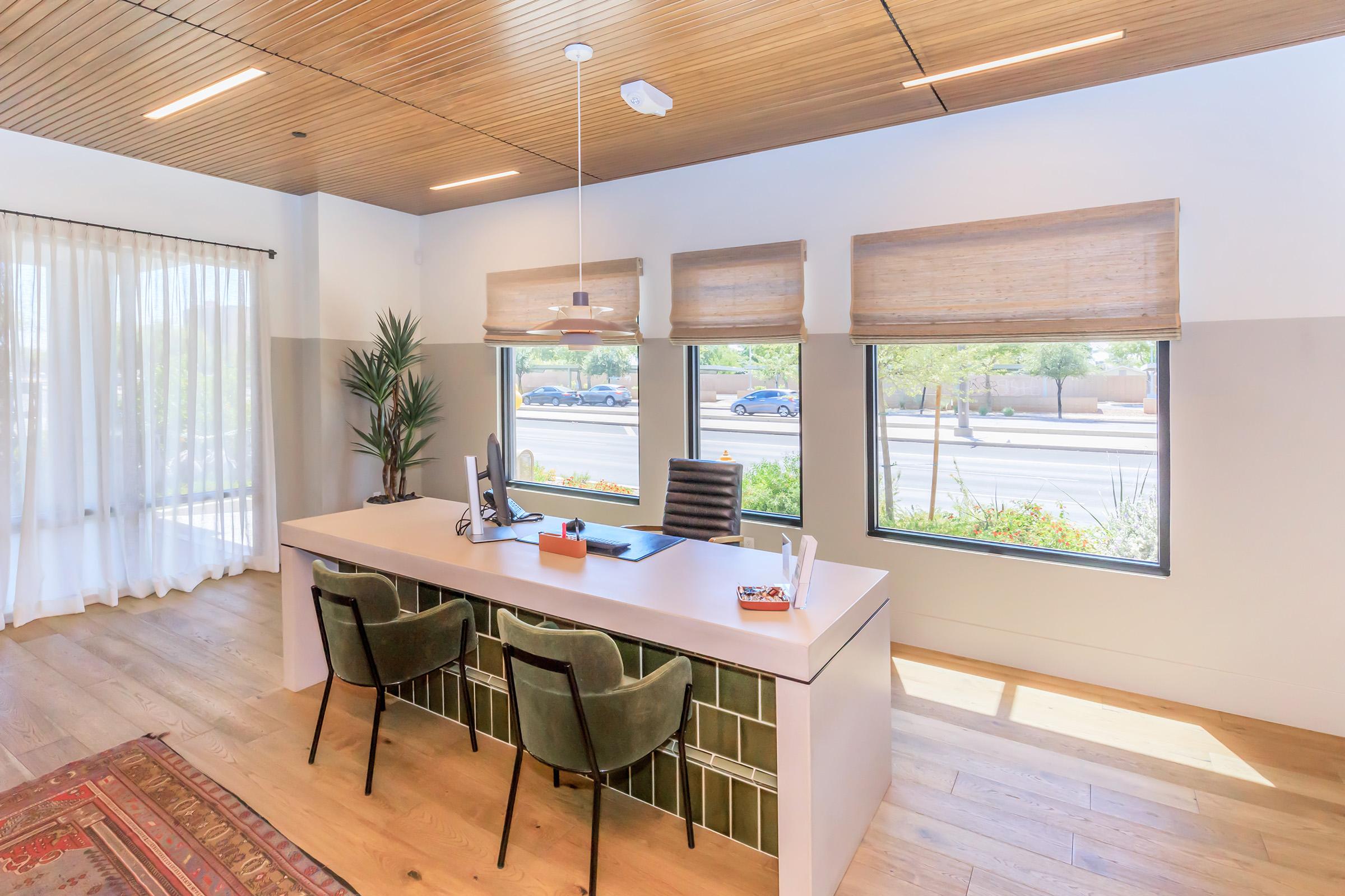 A modern office space featuring a sleek desk with a computer and chair, accented by a green plant. Large windows provide natural light, and the floors are wooden. Simple, stylish decor includes woven shades and a patterned rug, creating a warm and inviting atmosphere.