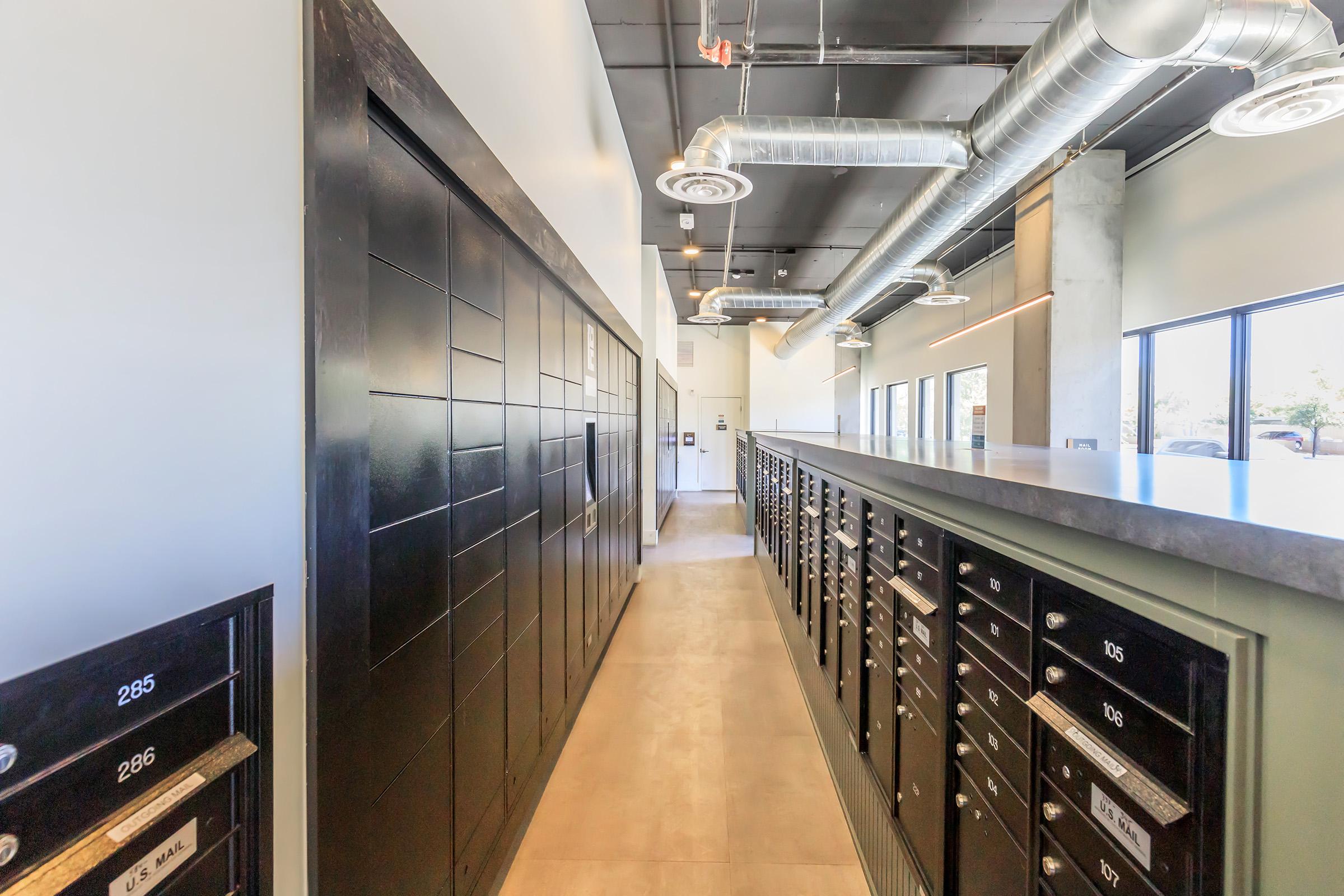 A modern hallway featuring black mailboxes along the left side, with a sleek counter on the right. The ceiling has exposed ducts and industrial lighting. Large windows allow natural light to fill the space, highlighting the contemporary design and clean lines of the interior.