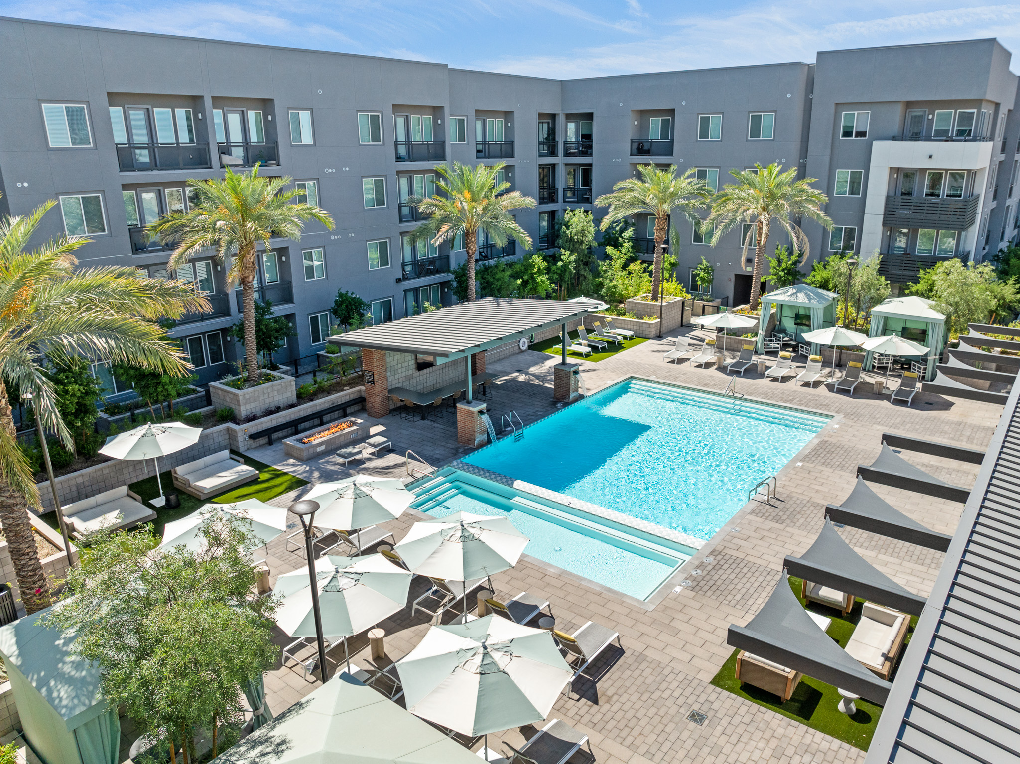 A spacious outdoor pool area surrounded by palm trees and lounge seating. Modern apartment buildings are in the background, with shaded cabanas and umbrellas providing relaxation spaces. Clear blue sky above enhances the inviting atmosphere of the pool area.