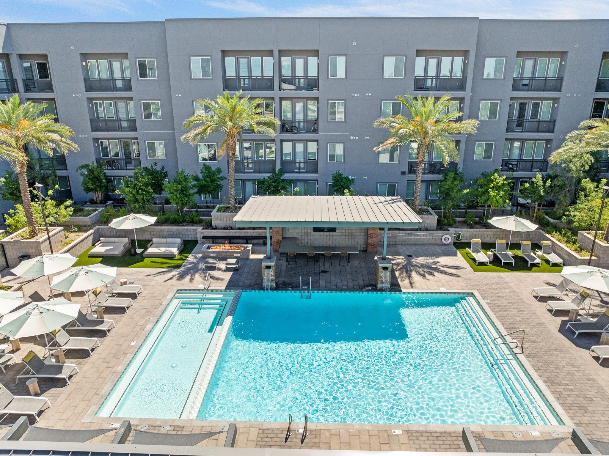Aerial view of a modern apartment complex featuring a large swimming pool surrounded by lounge chairs and dining areas. Palm trees provide shade, and a covered pavilion is situated nearby. The building in the background has multiple balconies and large windows, creating a vibrant outdoor living space.