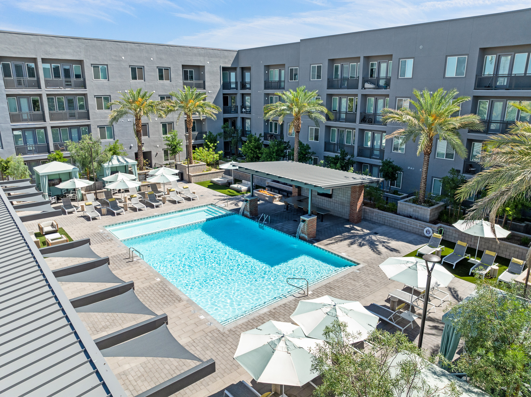 A crystal-clear swimming pool surrounded by lounge chairs and umbrellas, set within a contemporary apartment complex. Palm trees and landscaped greenery enhance the outdoor leisure area. The building features multiple balconies and large windows, providing a modern aesthetic against a blue sky.