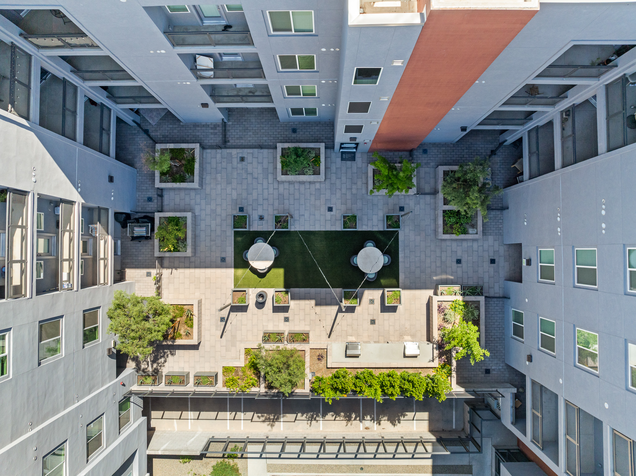 An aerial view of a modern urban courtyard surrounded by buildings. The space features green grass, trees, and landscaped areas with seating. Two large white umbrellas are set up, providing shade, and pathways connect different areas of the courtyard. The overall design is sleek and contemporary.