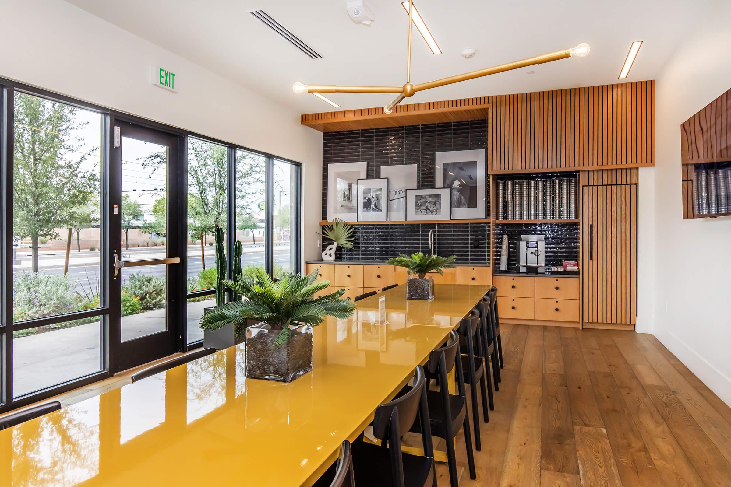 A modern meeting room featuring a large yellow table surrounded by black chairs. The room has large windows with views of greenery outside, wooden accents on the walls, and black-and-white photographs displayed. A coffee station is visible in the background.