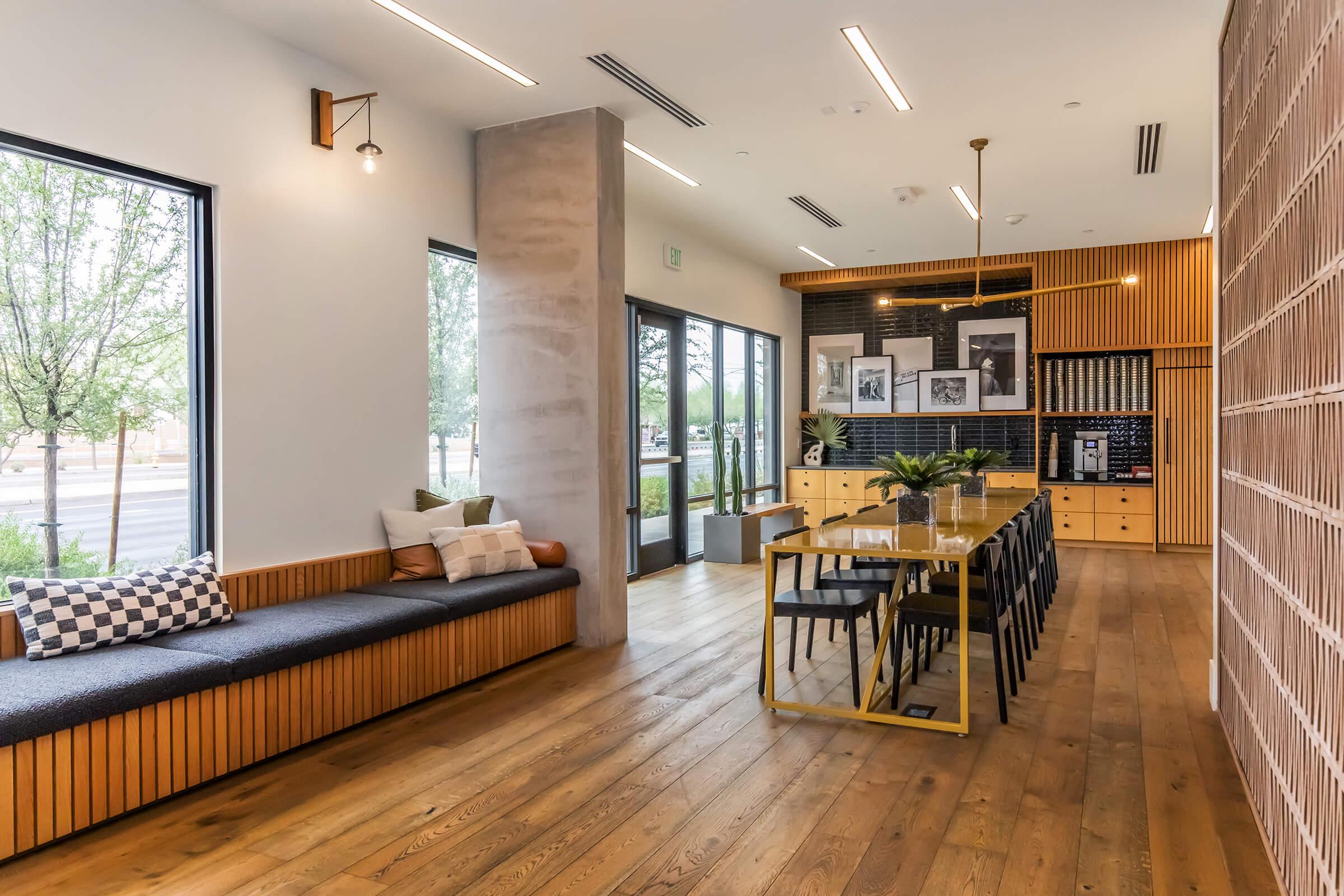 Interior of a modern space featuring a long bench with cushions, large windows, and a dining area with a wooden table and chairs. There is a wooden cabinet with framed photographs on the wall, and plants are placed throughout the room, creating a cozy and inviting atmosphere.