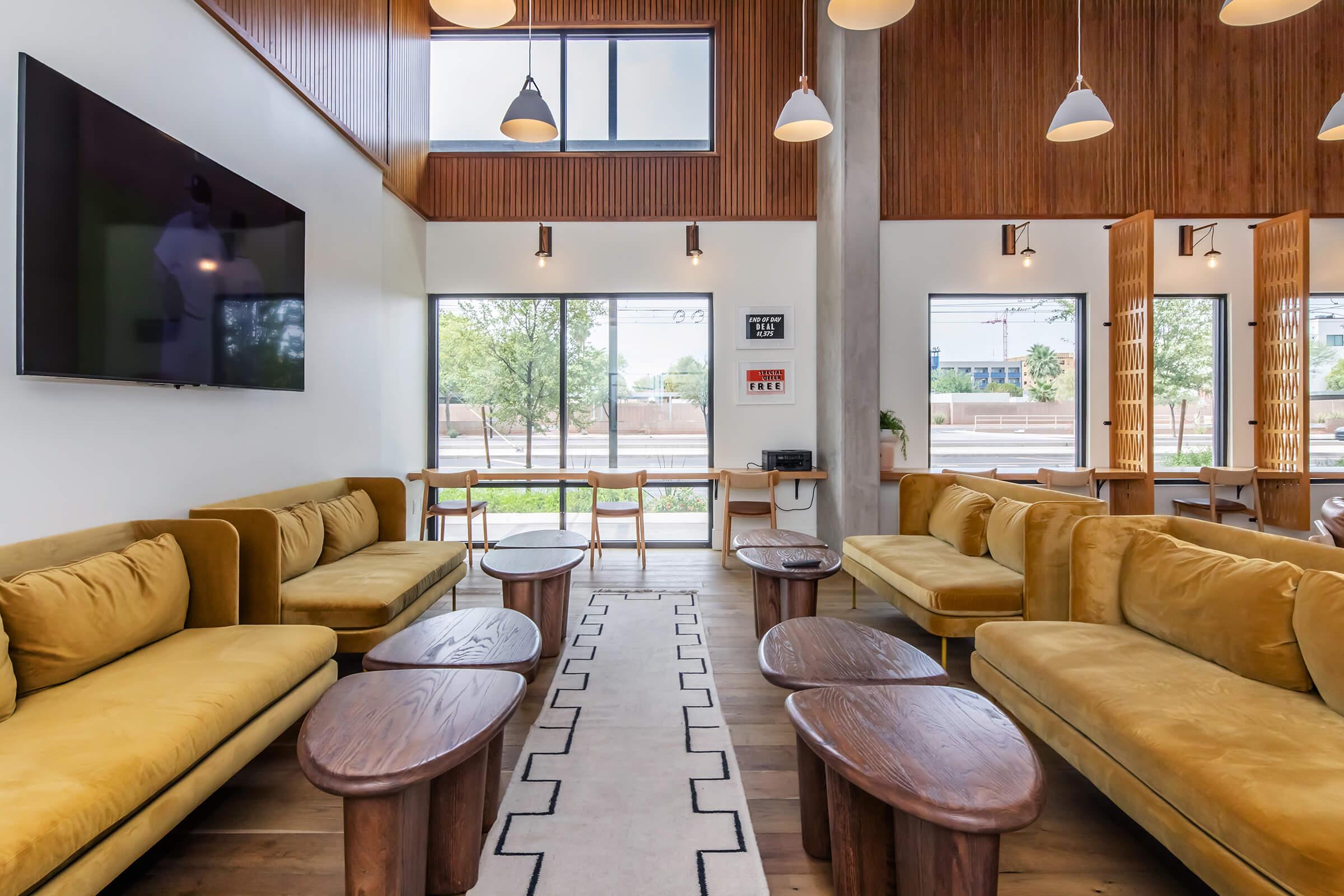 A modern lounge area featuring yellow sofas and wooden coffee tables, with large windows allowing natural light. The walls are adorned with wood paneling, and there is a television mounted on one wall. A minimalist design creates an inviting atmosphere, complemented by subtle lighting fixtures.