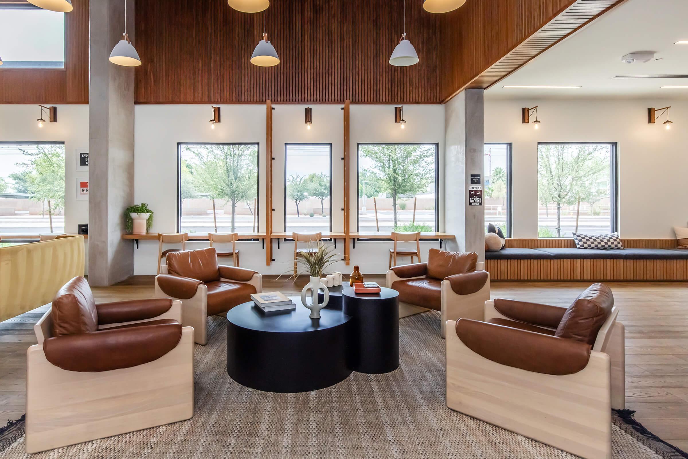 A modern lounge area featuring four brown leather chairs arranged around a black circular coffee table. The space is bright with large windows showcasing trees outside, and wooden accents in the ceiling and walls. A cozy atmosphere is created with a rug and decorative items on the table.