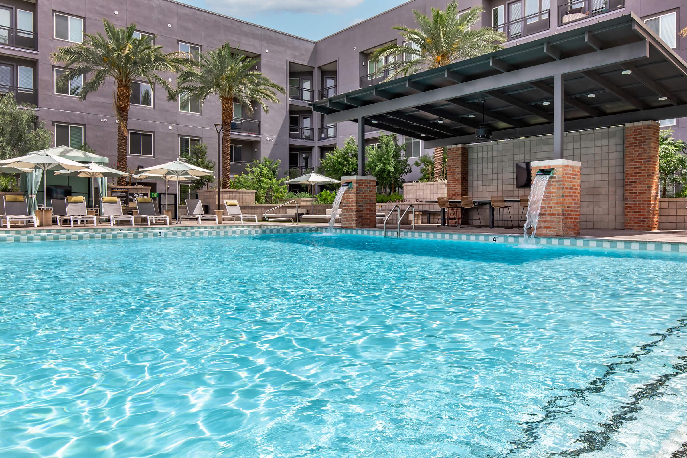 A clear swimming pool with rippling water and serene reflections, surrounded by lounge chairs and umbrellas. Palm trees provide shade in the background, with a modern building featuring large windows visible. A covered area with an outdoor grill enhances the inviting atmosphere.