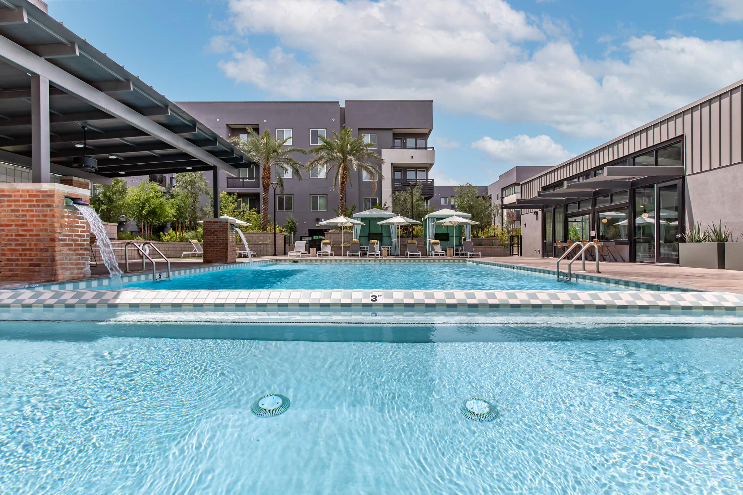 A clear blue swimming pool with a checkered border, surrounded by lounge chairs and green umbrellas. Palm trees and landscaped greenery are visible in the background, alongside modern buildings. Bright sunny sky with fluffy clouds above.