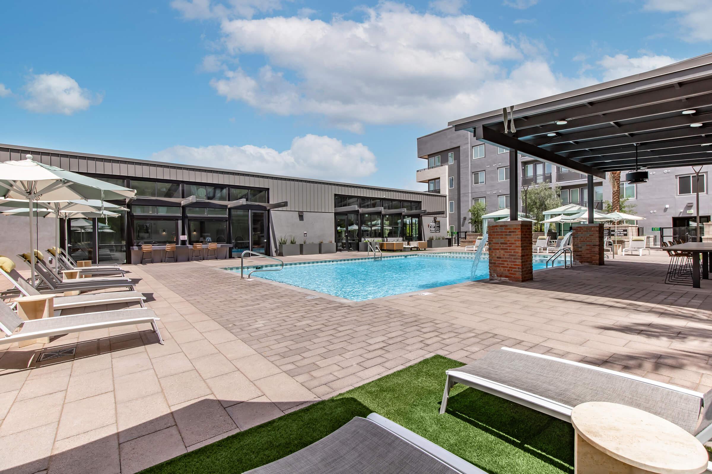 A modern outdoor pool area featuring a swimming pool surrounded by lounge chairs, shaded by umbrellas. In the background, there are contemporary buildings and a patio with tables, illuminated under a bright blue sky with a few clouds.
