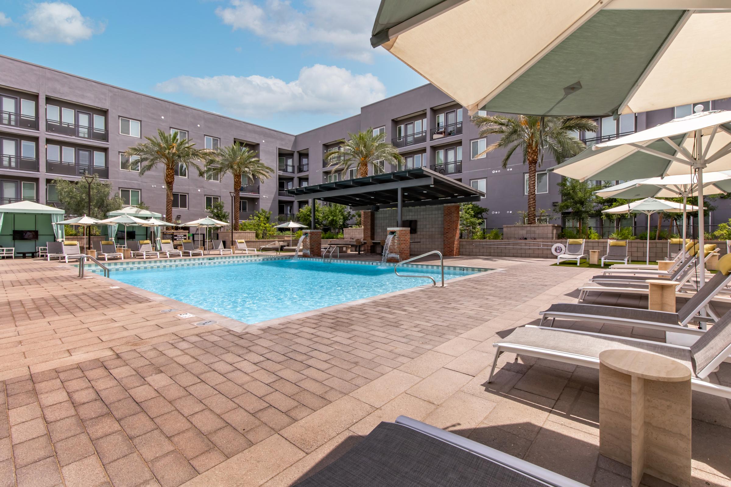 A sunny outdoor pool area featuring blue water, gray paving stones, and several lounge chairs. Umbrellas provide shade, and palm trees and greenery surround the pool. In the background, a modern apartment building is visible.