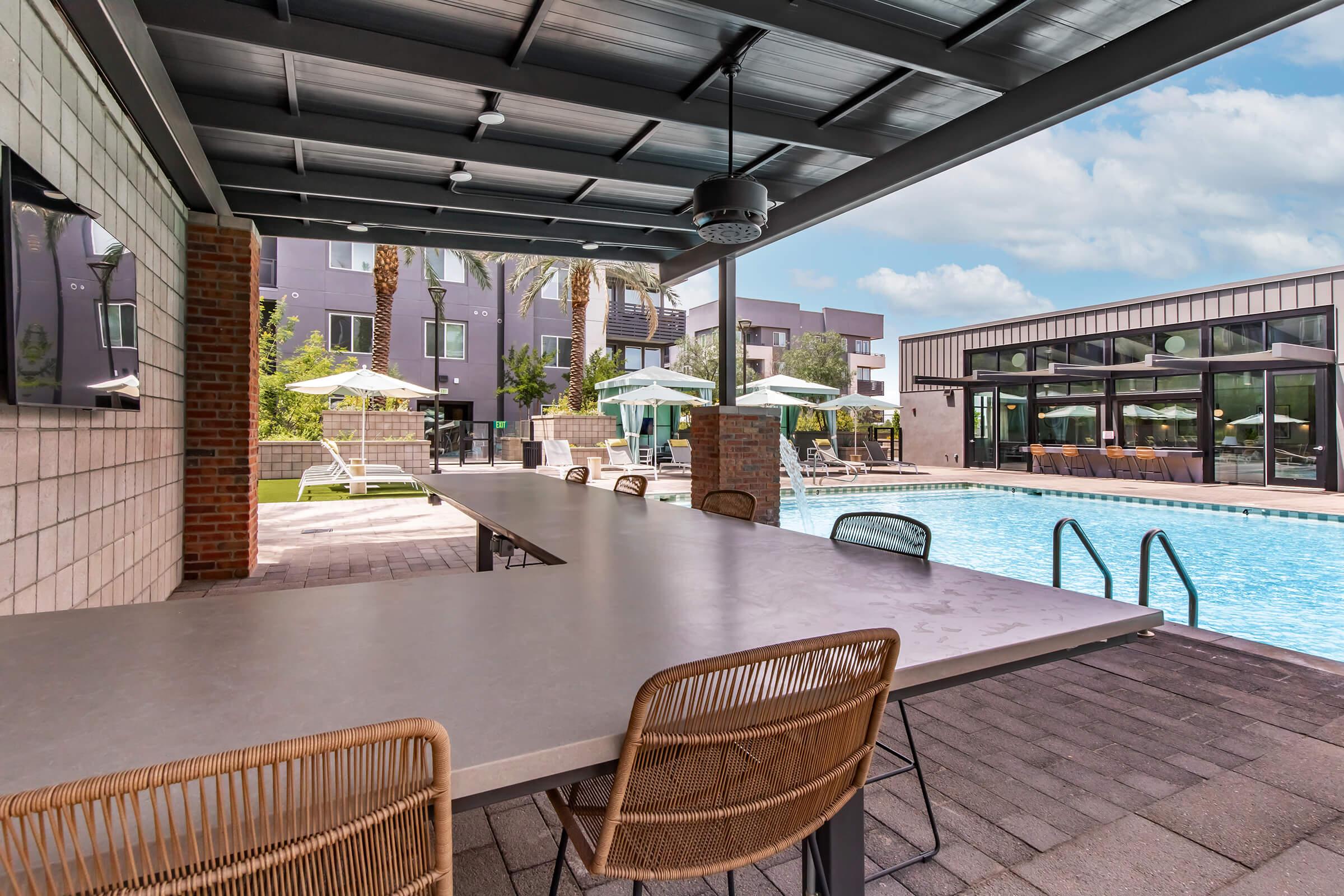 Outdoor pool area with a large, modern table and wicker chairs under a shaded pergola. In the background, there are lounge chairs and umbrellas beside the clear blue pool. Nearby buildings feature large windows, creating a relaxed, inviting atmosphere. Lush greenery and palm trees add to the ambiance.